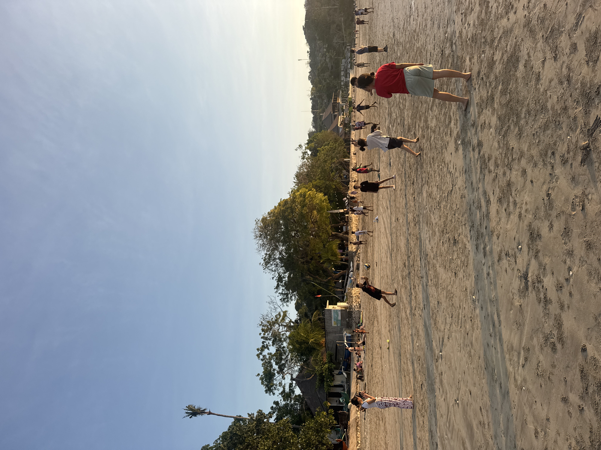 A wide shot captures a group of people playing volleyball on a sandy beach in Kuta Selatan, Indonesia. The sun is low in the sky, casting long shadows and bathing the scene in a warm, golden light. The beach is populated with individuals of various ages, engaged in recreational activities. Some are actively playing volleyball, while others are seated or standing in groups, observing or socializing. The background features lush green trees, some with palm fronds, and a few buildings with thatched roofs, suggesting a tropical, coastal resort area. The sky is a clear, pale blue, with a faint hint of haze near the horizon. The sand is a light brown, textured with footprints and small debris. The overall atmosphere is relaxed and lively, indicative of a typical day at a popular beach destination.