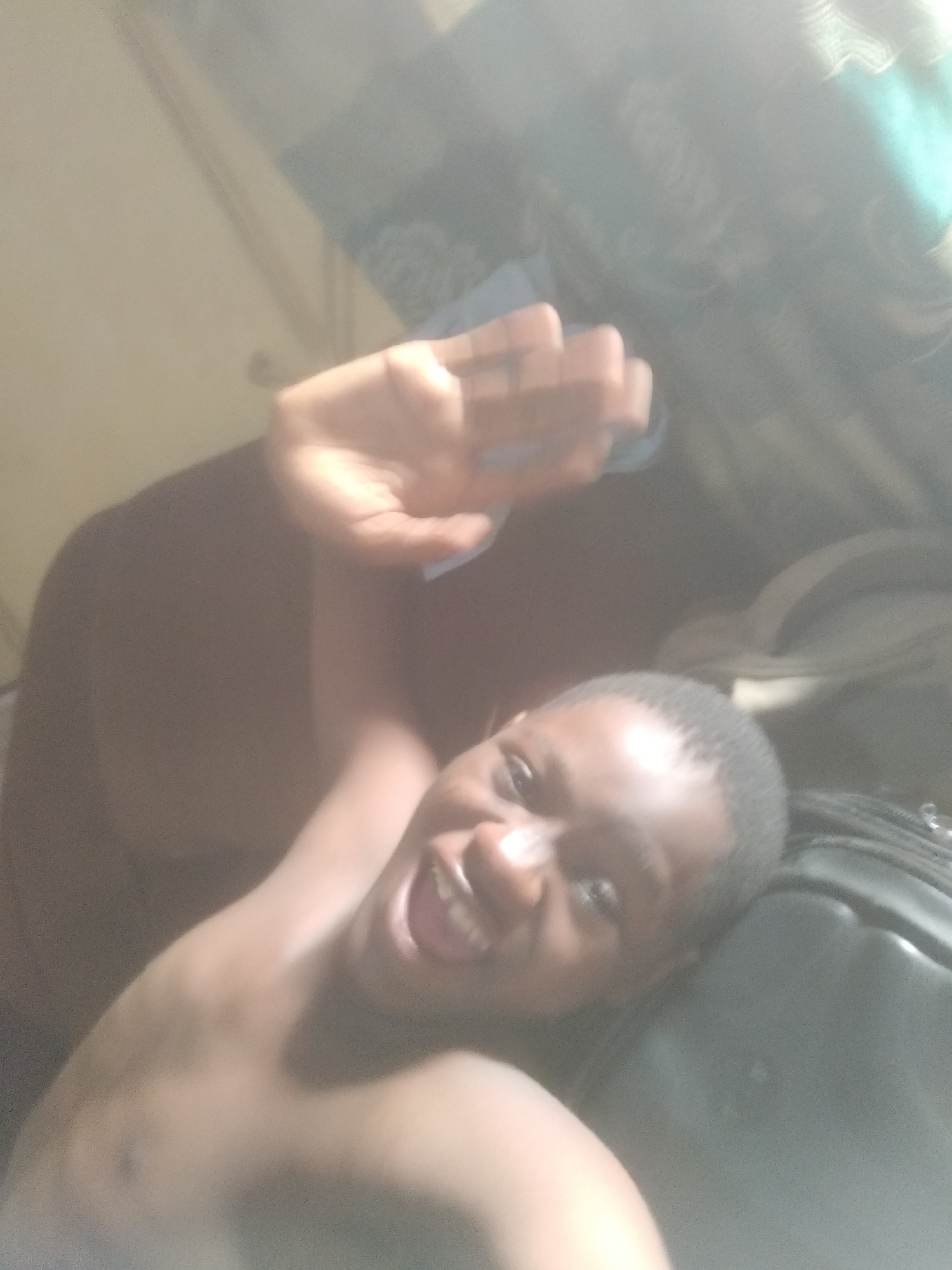 The image captures a young person, likely male, with dark skin and short dark hair, in an indoor setting, consistent with a bedroom or living area in Shangisha, Nigeria. The subject is lying down, positioned at an angle, and looks directly towards the camera with a wide, open-mouthed smile, conveying a strong sense of happiness and joy. Their left hand is raised and extended upwards, partially obscuring the top of the frame, possibly waving or shielding their eyes from a bright light source. The image has a somewhat hazy, diffused quality, suggesting bright daylight or a strong light source creating lens flare. A dark-colored piece of luggage or a bag is visible to the right of the subject's head. Faint, blurred patterns of fabric, possibly bedding or curtains, are discernible in the background. No visible text is present in the image.
