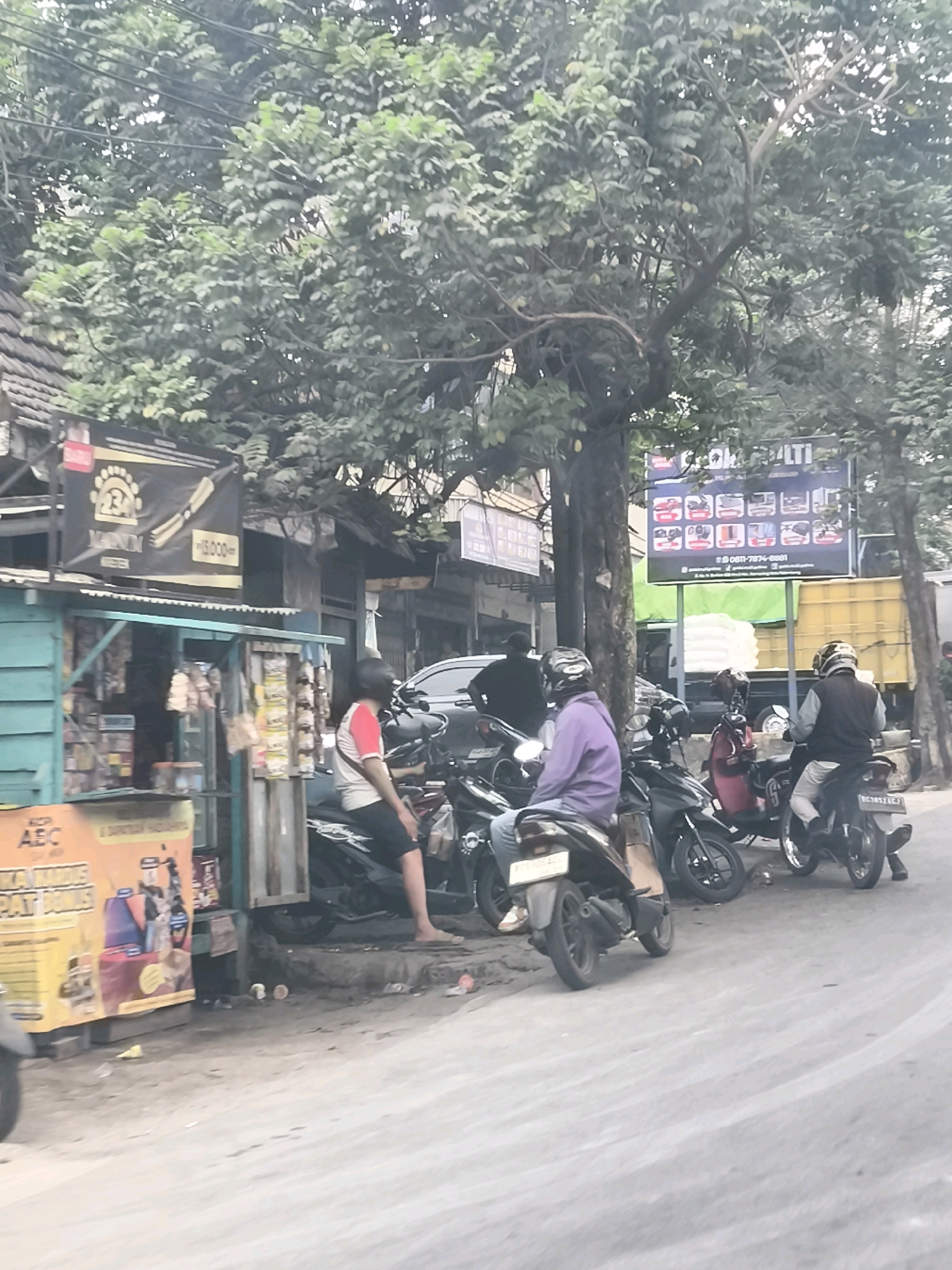 The image depicts a street scene in Palembang, Indonesia, likely in a commercial area given the presence of small shops and signage. A turquoise-colored kiosk, stocked with various goods, is visible on the left. Adjacent to it, a black banner advertising a product called "MAGNUM" with a price of "15.000" is displayed. Several individuals are present, most of them seated on motorcycles. One person is wearing a purple jacket and a helmet, while another wears a red and white shirt and black shorts, also on a motorcycle. The background shows more buildings, trees, and a billboard with multiple images and text in Indonesian. A yellow truck is parked in the background. The ground appears to be unpaved and dusty.