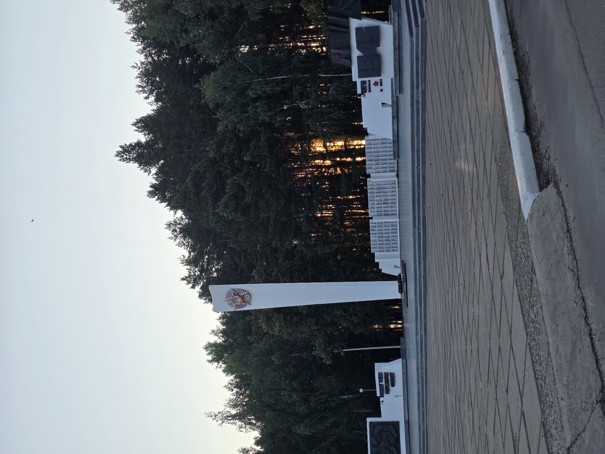 The image depicts the Memorial of Eternal Glory in the city of Chaikovsky, Russia. The memorial features a tall, white obelisk topped with a red and gold emblem. To the left of the obelisk, there is a section with a bas-relief and text in Cyrillic. To the right of the obelisk, there are rows of memorial plaques and large, dark numbers that appear to be "1941-1945". The memorial is situated in a paved square, with a line of trees forming a backdrop. The sun is setting, casting a warm glow through the trees. The sky is a pale blue, with a small bird visible in the upper portion of the image. The foreground shows the edge of the paved area and a road.