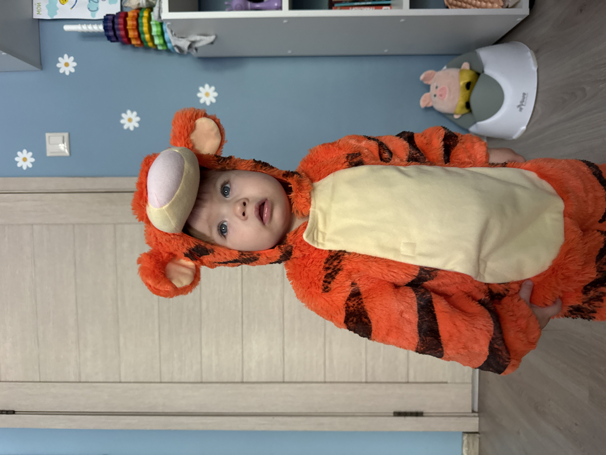 A young child, appearing to be around 1-2 years old, stands in a dimly lit room, dressed in an orange and black Tigger costume. The child has blue eyes and is looking up and to the right, with their mouth slightly open. The costume has large, fuzzy ears and a beige belly. In the background, a white door with horizontal paneling is visible to the left. To the right, a light blue wall is decorated with several white daisy stickers and a light switch. Further to the right, a white shelf unit stands with various items, including a stack of colorful rings and a pink pig toy sitting on a potty. The floor appears to be light-colored wood laminate. The overall environment suggests a nursery or playroom. There is no information to suggest the location is in Moscow, Russia.