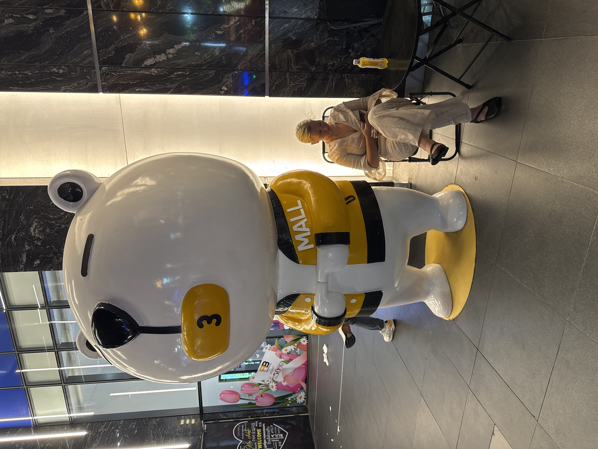 A life-sized cartoon bear sculpture, white with a yellow jacket and a yellow oval on its chest with the number "3" in black, stands on a yellow circular base in front of a large marble-patterned pillar. A blonde woman with light-colored clothing is sitting on a folding chair to the right of the sculpture, looking at her phone. She is seated at a small black table with a bottle of yellow liquid. In the background, a banner with images of tulips and text about a sale is visible. The flooring is composed of dark grey square tiles. The scene appears to be indoors, possibly in a shopping mall or public area. The location is Nha Trang Ward, Vietnam.