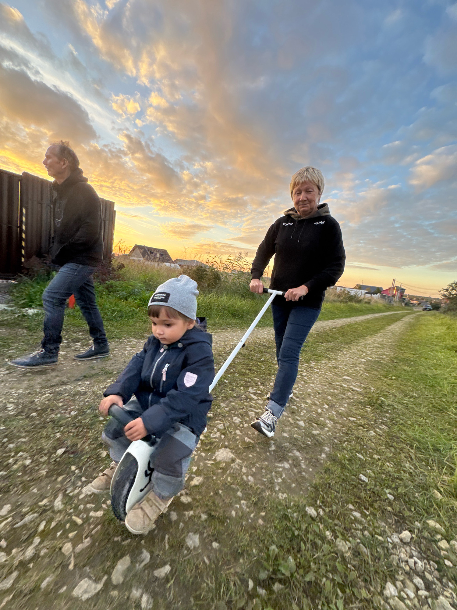 In Kempelevo, Russia, a low-angle medium shot captures a rural scene at dusk. A young child wearing a gray beanie and a dark blue jacket is seated on a white and black electric unicycle, holding onto its handlebars. To the right, a woman with short blonde hair, wearing a black hoodie and blue jeans, is holding a white handle attached to the unicycle, appearing to guide the child. Further in the background on the left, a man in a dark hoodie and jeans walks away from the camera, looking towards the left. The ground is a dirt path with patches of grass and scattered stones. In the background, a wooden fence, houses with roofs, and trees are visible. The sky is filled with dramatic clouds illuminated by the setting sun, casting a warm orange and yellow glow. The overall mood is serene and familial, set against the backdrop of a natural landscape.