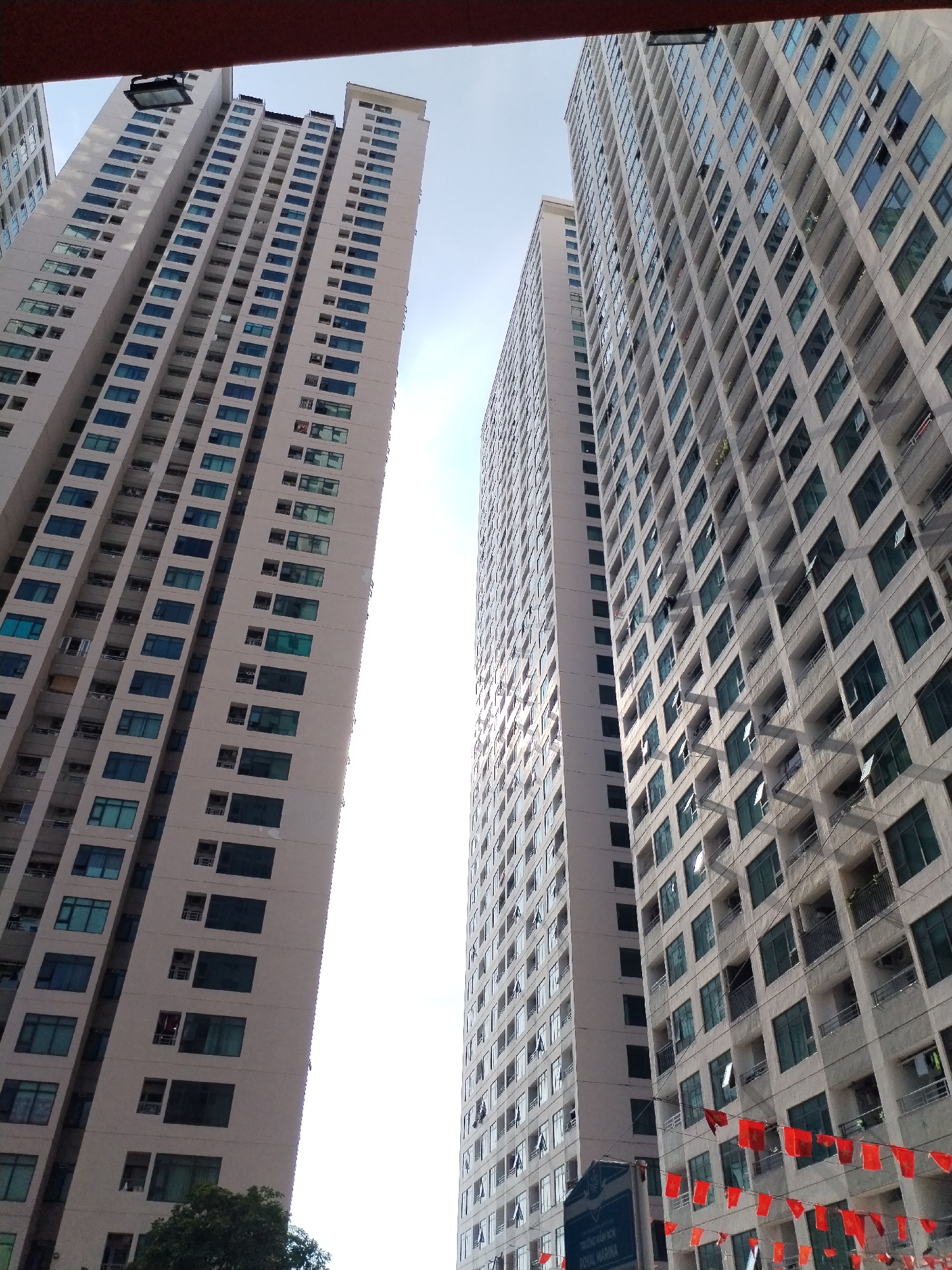 The image shows two tall, modern apartment buildings in Bac Nha Trang Ward, Vietnam. The building on the left is lighter in color and has a uniform pattern of windows. The building on the right has a more varied facade with balconies and air conditioning units visible. Both buildings are very tall, reaching high into the sky. There are strings of red flags hanging across the front of the building on the right, suggesting a festive occasion. A street sign is visible at the bottom right of the frame, with text that is difficult to read but appears to say "ROYAL MARINA." The sky is bright and clear, indicating a sunny day. The angle of the photograph is looking up at the buildings, emphasizing their height and scale. No people are clearly visible in the image.