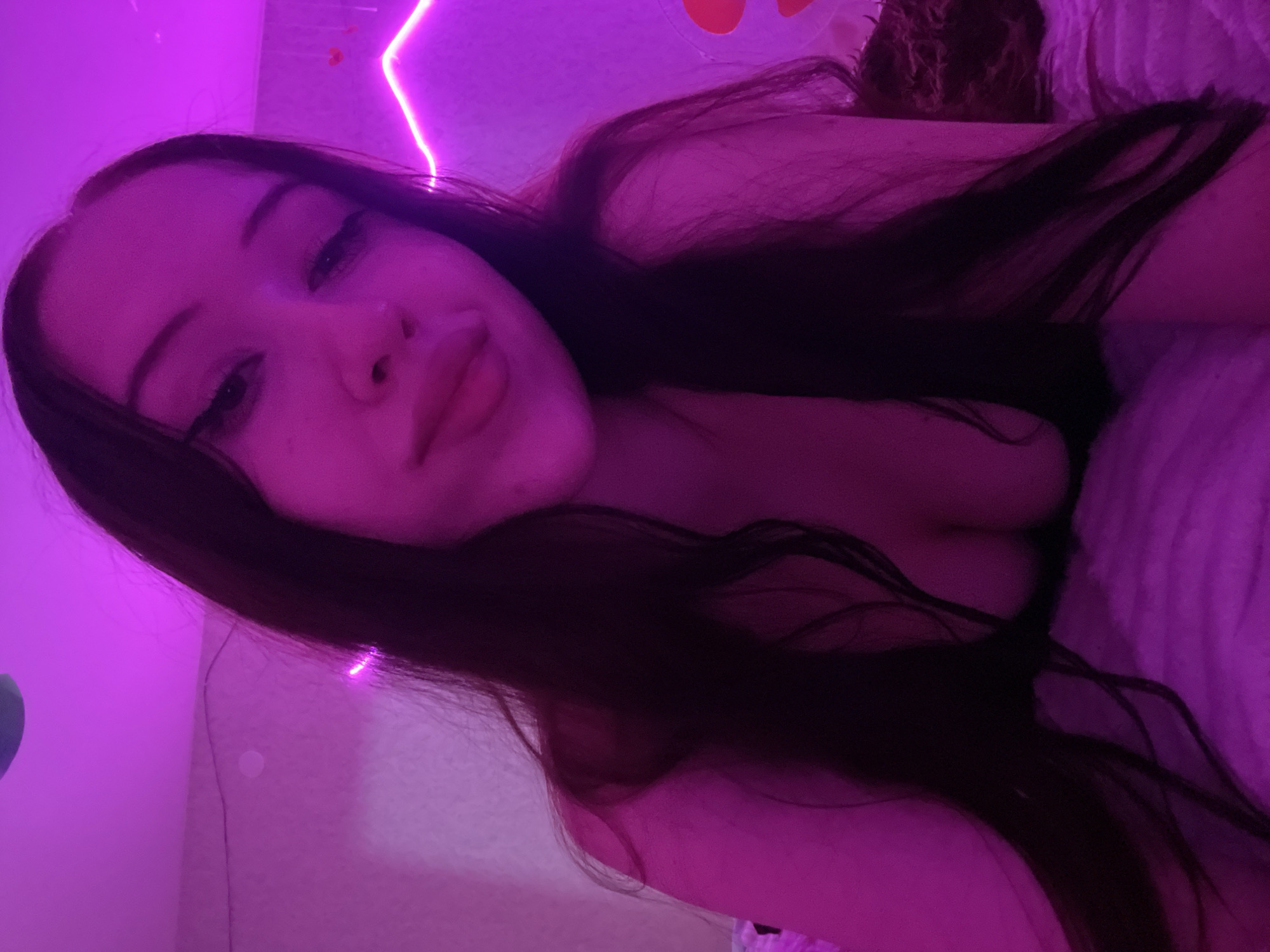 The image depicts a young woman in a dimly lit room, illuminated by pink and purple neon lights. She has long, dark hair and is wearing a dark-colored top. Her eyes are looking towards the camera with winged eyeliner, and her lips are slightly parted. The background features a wall with neon lighting, including a decorative heart shape. The setting appears to be a bedroom, with a bed covered in a textured pink blanket visible in the foreground. The overall ambiance is intimate and artistic, with the neon lighting casting a vibrant glow on the subject and her surroundings. The image does not contain any elements that would allow for the identification of its specific location within the city of Votkinsk, Russia.