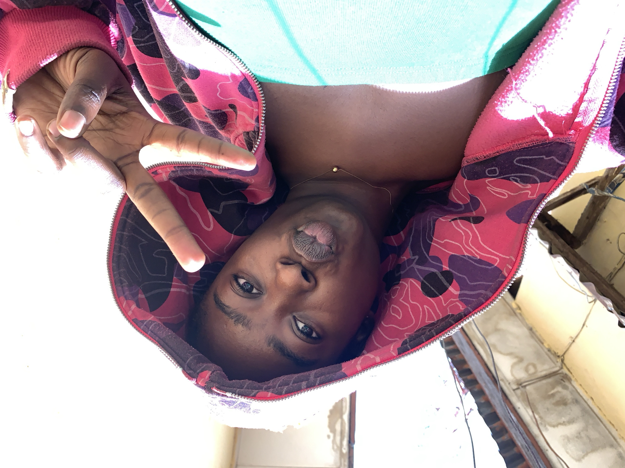 A young individual is depicted in a selfie, appearing to be located outdoors in Rafin Par, Nigeria, as suggested by the architectural features present. The person is wearing a pink hooded jacket with a distinctive pattern of abstract shapes in darker shades. A light green shirt is visible underneath the jacket. A thin gold chain necklace adorns the neck. The individual is holding up their hand in a peace sign. The background features a building with a corrugated iron roof and external electrical wiring, likely within an urban setting. The overall lighting suggests it is daytime.