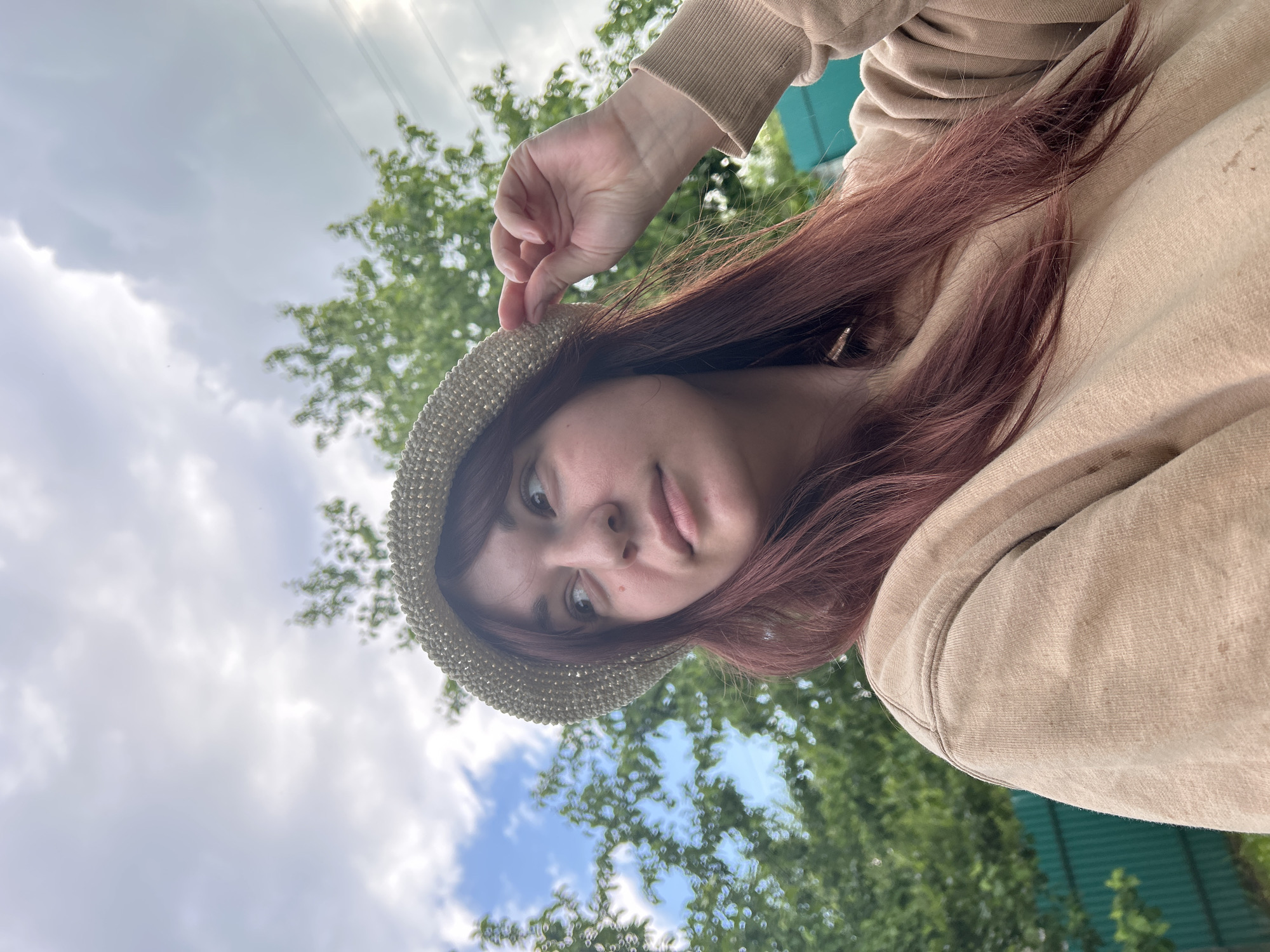 A young woman is featured in a close-up, low-angle photograph, which is rotated. She looks directly at the viewer with a calm, neutral expression. Her long, reddish-brown hair frames her face and falls onto her shoulders and chest. She is wearing a light beige or tan hooded sweatshirt. Her right hand is visible, gently holding the brim of a light-colored, possibly crocheted or woven, hat on her head.
The background shows an outdoor setting under a partly cloudy sky with patches of blue. Lush green tree foliage, mostly out of focus, is visible around her. A section of a green corrugated metal fence or structure can be seen behind her right shoulder in the background. Faint lines, possibly power lines, are present against the sky in the upper left. The image was taken during the daytime in what appears to be a natural or semi-natural environment, located in Фоки, Russia. There is no visible text in the media file.