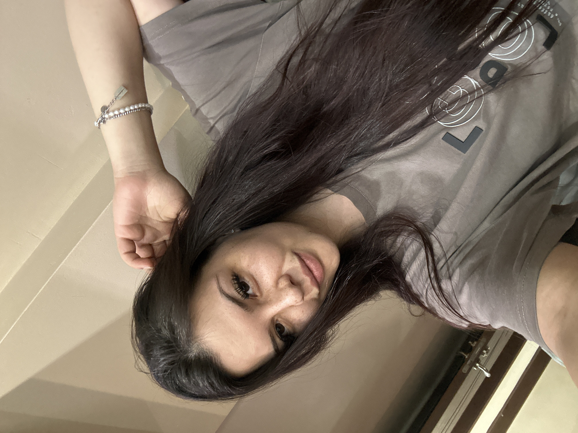 The image is a selfie taken by a young woman with long, dark brown hair. She is wearing a grey t-shirt. Her left hand is resting on her head, and she is wearing a bracelet on her left wrist. Her right arm is visible, and she is leaning on it. The background appears to be an indoor setting with a beige wall. There is no discernible indication of the city of Sliven, Bulgaria, or any specific urban environment.