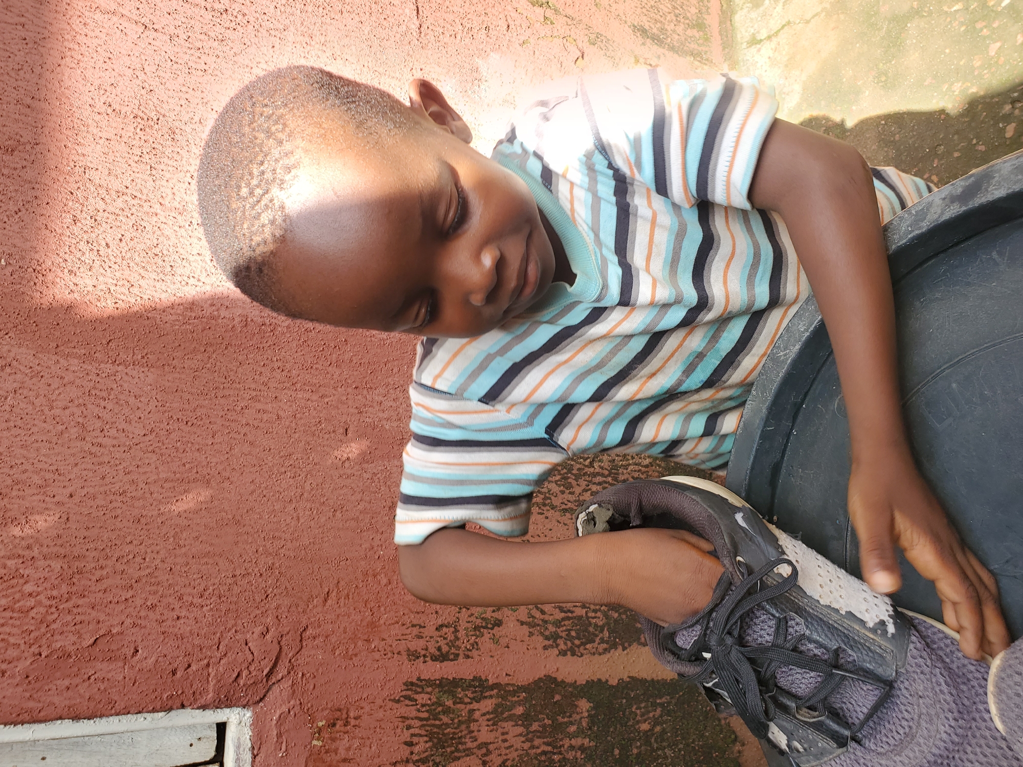 This media file shows a young boy resting against a textured, reddish-brown wall. He is wearing a striped t-shirt with horizontal bands of blue, black, white, and orange. His eyes are closed, and he appears to be sleeping or deep in thought. His right arm rests on a dark, textured surface, likely a large container, while his left hand is holding a worn black and white sneaker. The lighting suggests it is daytime, possibly late afternoon, with sunlight casting shadows. The setting appears to be outdoors, with hints of a rough concrete or paved ground in the background. The overall mood is peaceful and relaxed. The image was taken in Jos, Nigeria. There is no visible text in the image.