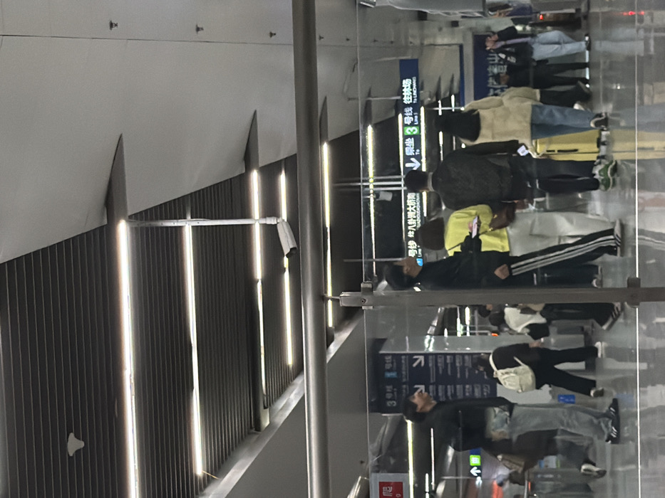 The scene depicts a concourse within Nanjing South Railway Station, located in Yuhuatai District, Nanjing, China. The environment is a brightly lit, modern indoor space with a ribbed metal ceiling, integrated linear LED lighting, and large white ceiling panels. A white surveillance camera is mounted from the ceiling, positioned to view the area below. Horizontal metal railings and vertical transparent glass panels serve as barriers. Signage in simplified Chinese and English is visible, indicating directions for "3号线 往林场" (Line 3 To Linchang) and "1号线 往八卦洲大桥南" (Line 1 To Baguazhou Bridge South).
Approximately fifteen individuals are present, all appearing to be adults, engaged in transit. In the mid-ground, a group of four people walks away from the viewer. From left to right within this group: a male in a black tracksuit with white side stripes, holding a light-colored object; a female in a bright yellow top and light-colored trousers, carrying a brown bag; a male in a dark, possibly patterned, jacket and dark trousers; and a female in a light beige fluffy jacket and blue jeans, pulling a yellow wheeled suitcase. To the far left, a male wearing a black hoodie, light blue jeans, and Adidas sneakers is positioned facing right, exhibiting an open-mouthed expression. In the background, several other individuals are visible walking or standing near what appear to be entry or exit points, one wearing a white backpack. The floor exhibits reflections of the overhead lighting and figures.