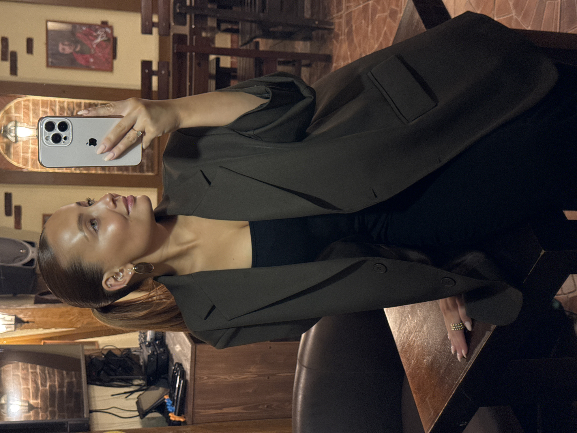 A woman with her hair pulled back in a ponytail is taking a selfie in a dimly lit room. She is wearing a dark blazer over a black top and has a ring on her right hand. She is holding a smartphone in her right hand, which is angled to capture her reflection in a mirror. The background shows a brick wall, wooden furniture, and a framed picture of a man in a red garment. The scene appears to be a restaurant or bar.