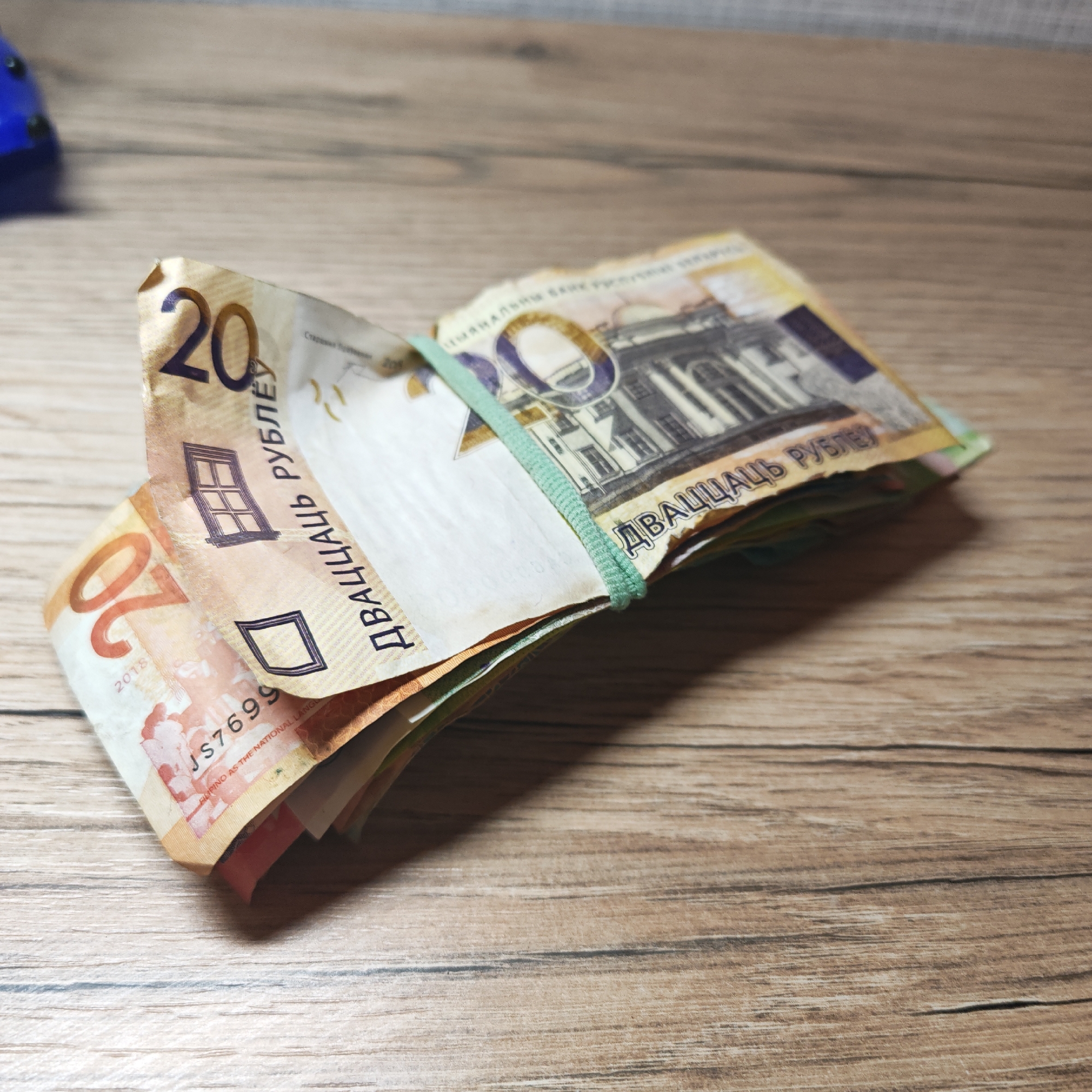 The image shows a stack of Belarusian 20 ruble banknotes, secured with a green rubber band. The banknotes are folded and the top note displays the number "20" prominently. The text "ДВАЦЦАЦЬ РУБЛЕЎ" (Twenty Rubles) is visible. The serial number "JS7699" is also visible. The banknotes are lying on a wooden surface with a light brown, wood-grain pattern. Part of a blue object is visible in the background, upper left corner. The scene does not appear to be in the city of Rostov, Russia.