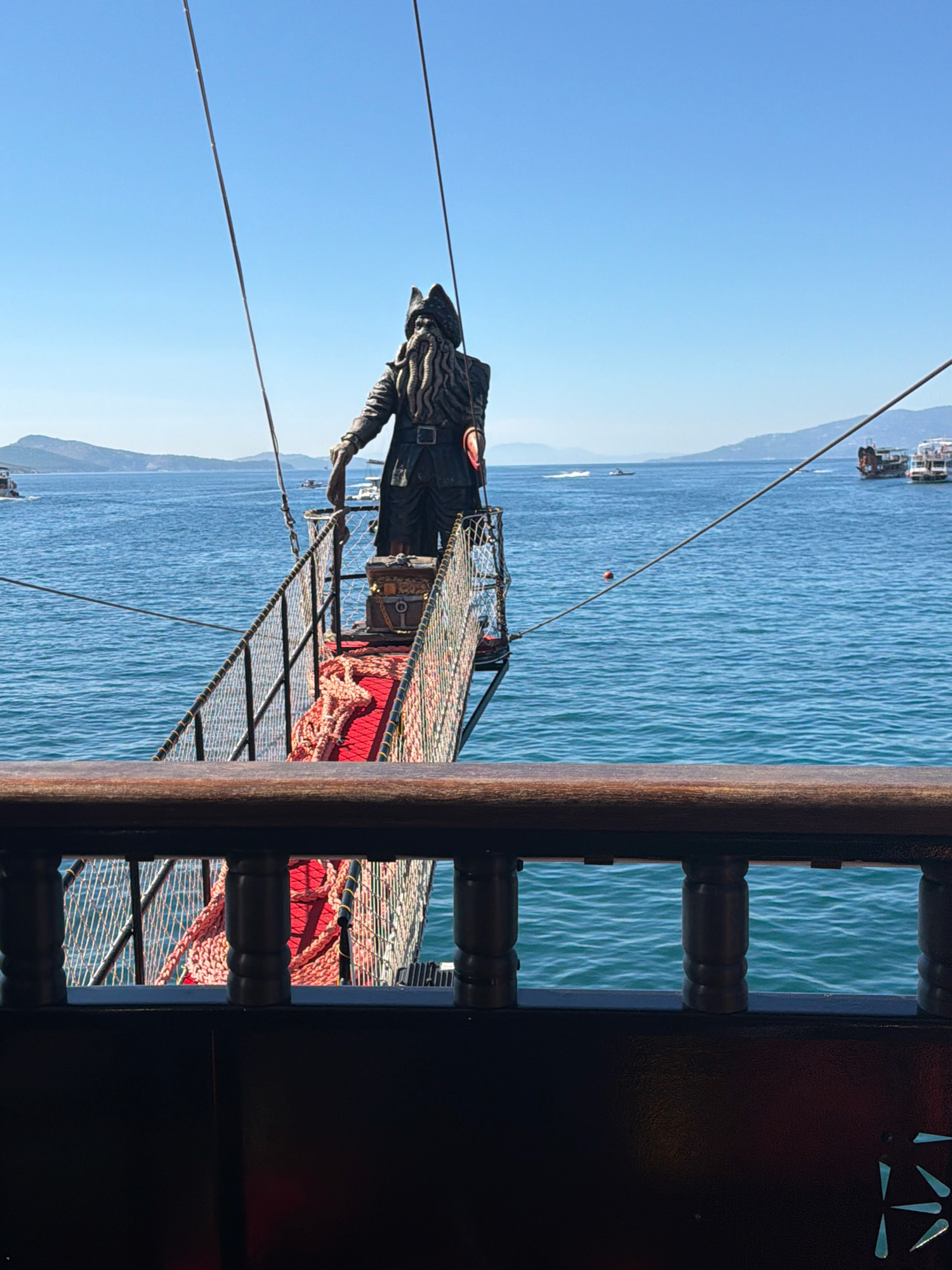 A statue of a pirate with tentacles for a beard stands on a wooden platform extending over the blue sea. The pirate wears a black coat, a tricorn hat, and a belt. A red carpet and thick ropes are laid out on the platform, and a treasure chest sits near the pirate's feet. The statue is positioned on a gangplank attached to a wooden ship, with the railing of the ship visible in the foreground. In the background, several other boats are scattered across the water, and a hilly coastline with some buildings is visible under a clear blue sky. This scene is likely located in the city of Saranda, Albania, known for its scenic coastline and boat tours.