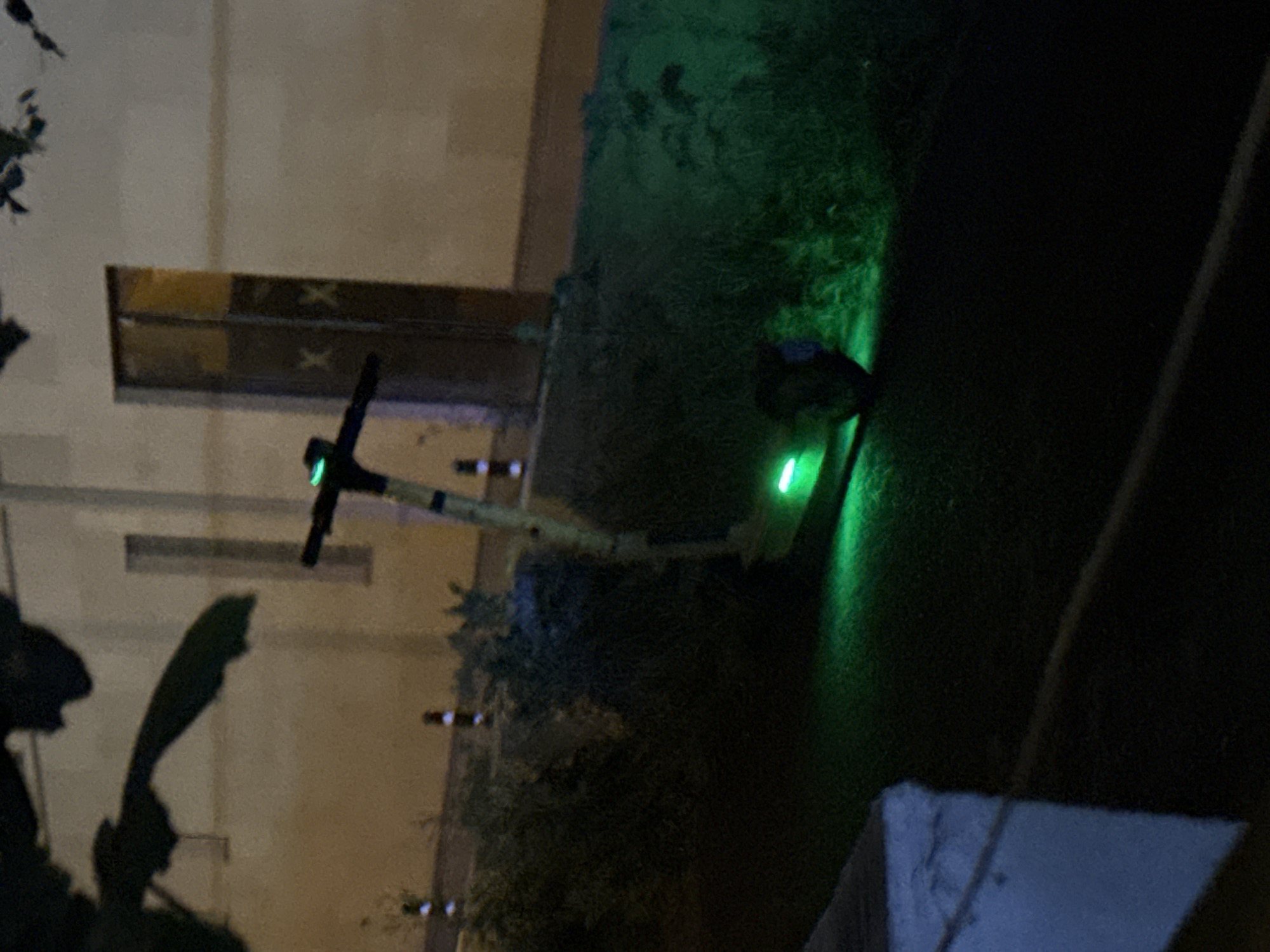 The image shows a street scene at night in Tbilisi, Georgia. In the foreground, a scooter with green LED lights is parked on a grassy area next to a paved surface. The scooter is silver and black with a green light illuminated at the base of the handlebars and on the rear wheel. The background features a light-colored building with a dark doorway that has two "X" marks on it. There are also several small, reflective bollards lining the edge of the grassy area. Leaves from a tree are visible in the upper left corner of the image. The overall lighting suggests it is nighttime or dusk, with artificial lights illuminating parts of the scene.