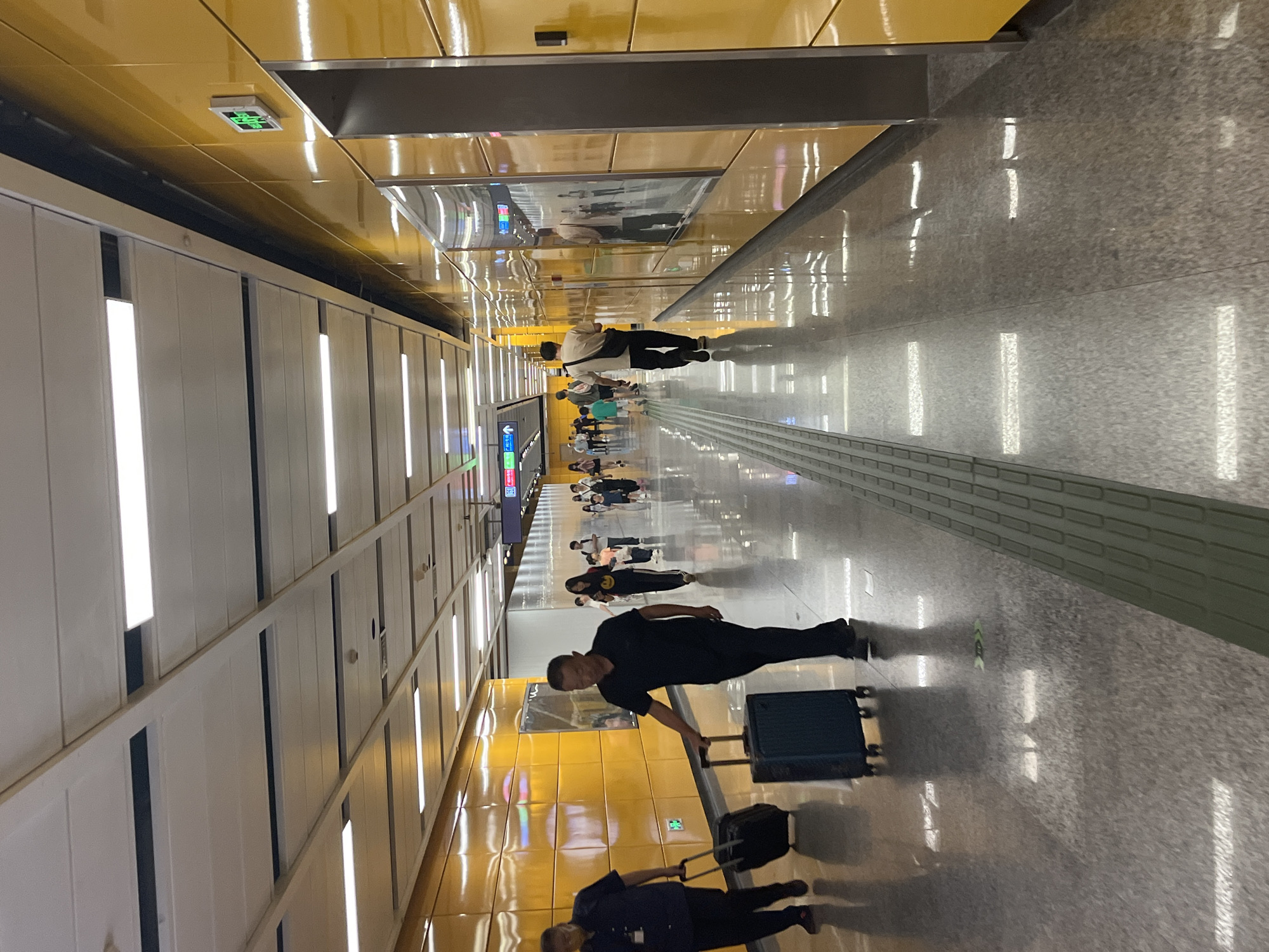 The image depicts a brightly lit, modern underground subway station in Panyu District, China. The walls are a vibrant yellow, with reflective surfaces that mirror the overhead fluorescent lighting. The floor is a polished grey granite, also highly reflective, showing streaks of light. In the foreground, two men are walking with rolling suitcases. The man on the left wears a dark blue shirt and pants, and is pulling a black suitcase. The man on the right wears a black polo shirt and dark pants, and is pulling a teal suitcase. Further down the corridor, a procession of people, including women, men, and children, are walking in the same direction. Some are carrying bags. Overhead, a grid of white rectangular lights illuminates the passageway. A green illuminated sign indicating an exit is visible on the right wall, and directional signs are present further down the tunnel. The overall impression is one of a clean, efficient, and busy public transportation hub.