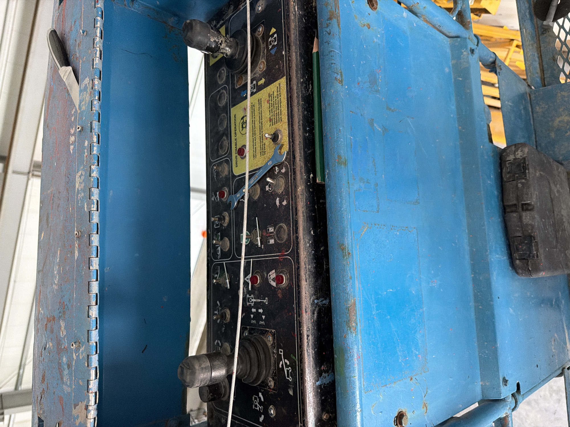 The image displays a close-up view of a blue control panel on a piece of machinery, likely a lift or aerial work platform. The panel is black with various buttons, switches, joysticks, and labels. Some of the labels are in yellow and describe operation procedures, such as "DRIVE ENABLE OPERATION." A green pencil is positioned horizontally across the lower portion of the panel, and a small metal wrench is also visible on the panel, resting on some of the controls. The surrounding machinery is painted blue and appears to be weathered, with some rust visible on the upper section of the panel and on the blue housing. The background shows the interior of a building, with white corrugated metal walls and ceiling structures, suggesting an industrial or workshop environment. The location is specified as Janowice, Poland, although there are no visual cues within the image that directly confirm this specific geographical location.
