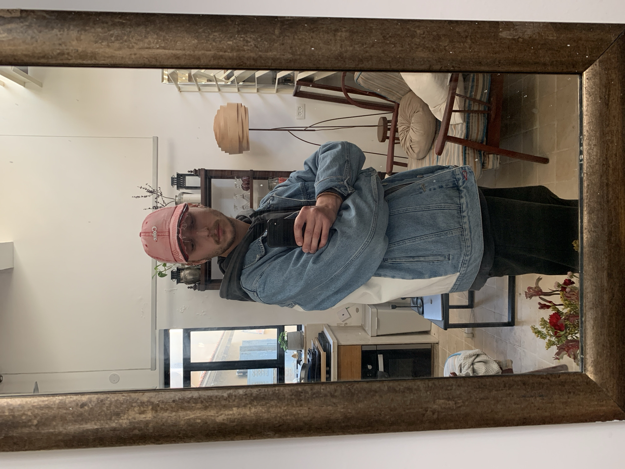 An individual, identified as a young adult male, is depicted in a mirror selfie within an indoor residential setting in Tel-Aviv, Israel. The male subject wears a reversed pink baseball cap with "Coke" branding visible, a dark grey hooded sweatshirt, a blue and white denim jacket, and dark trousers. His arms are crossed at chest level, holding a black smartphone with a protective case, angled to capture his reflection. Facial features include a mustache and a short beard; a silver-colored earring is visible in his left ear.
The reflected environment is a residential space with white walls displaying visible areas of paint damage, including peeling and discoloration, particularly on upper sections. The floor is covered with light-toned square ceramic tiles.
On the left side of the reflection, a kitchen area is observed through a multi-pane window. A small potted plant sits on the windowsill. Below, a countertop holds stacked dark baking trays. A light-colored wooden cabinet and a dark built-in oven are visible underneath, adjacent to a light grey waste bin. The exterior view through this window shows a building with light-colored walls and a narrow passage.
On the right side of the reflection, a living area is present. A tall, dark wooden shelving unit contains a lantern-style object, dried plant material in a vase, and multiple glass vessels. A floor lamp featuring a stacked, layered wooden shade and a dark metal base stands in front of a wooden armchair upholstered with striped fabric and featuring a round cushion. To the right of the armchair, another window with horizontal mullions reveals an exterior building. A dark, rectangular side table supporting a closed laptop and two black candlesticks is partially visible in the mid-ground. In the lower foreground of the mirror, a vase containing fresh cut flowers, including red and pink blooms with green foliage, is visible.