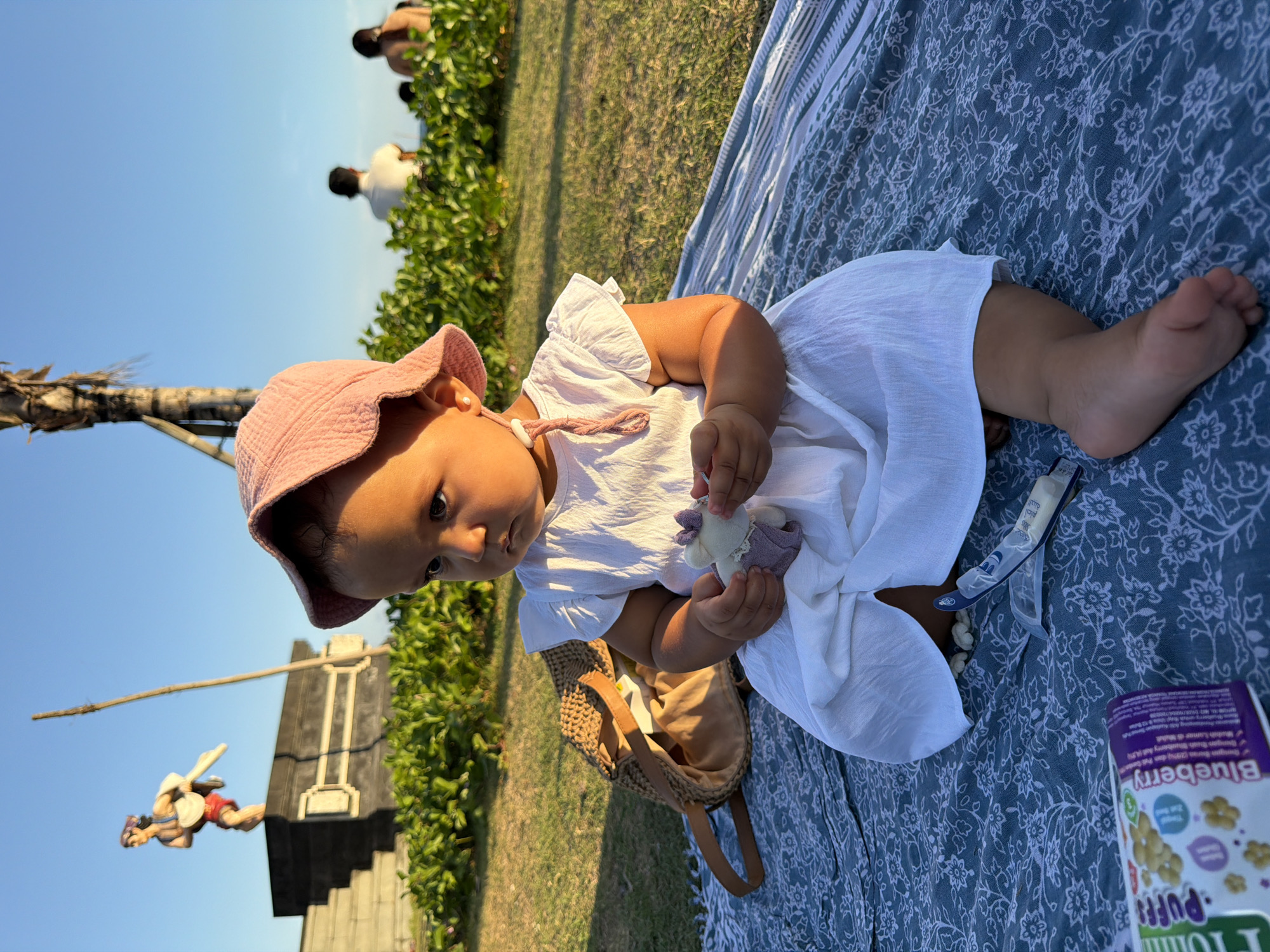 A young child wearing a pink hat and a white dress sits on a patterned blanket on a grassy area. The child is holding a small stuffed toy. In the background, a statue of a man is visible on a raised platform, and further back, several people are walking on a beach. Lush green bushes line the area between the child and the beach. The sky is clear and blue, and the sun is casting long shadows, indicating it is late afternoon or early evening. A woven bag and a box of baby puffs are placed next to the child on the blanket. The location appears to be a public park or beach area in Mengwi, Indonesia, possibly near Tanah Lot temple, given the statue and the coastal environment.
