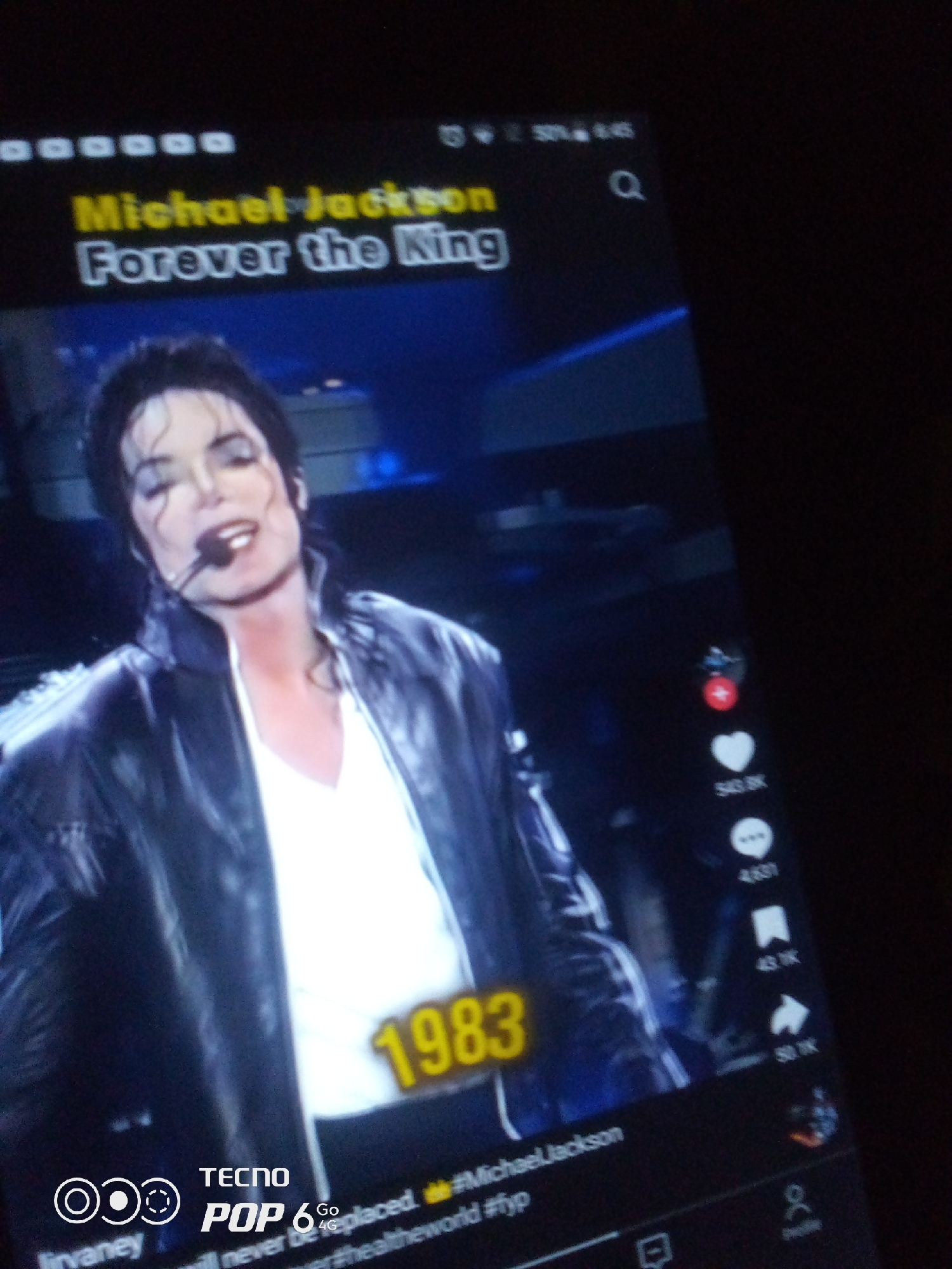 The image displays a screen, likely a smartphone or tablet, with a video playing. The video features a man, identifiable as Michael Jackson, wearing a black leather jacket over a white shirt. He is singing into a microphone. Text overlays on the screen read "Michael Jackson Forever the King" and "1983". The bottom of the screen shows app branding that includes "TECNO POP 6 Go". The user interface elements on the right suggest a social media platform, with icons for likes, comments, shares, and profiles. The lighting is dim, indicating the device is being viewed in a dark environment. There is no visible indication of the city of Howayi, Nigeria, or any other specific geographic location.