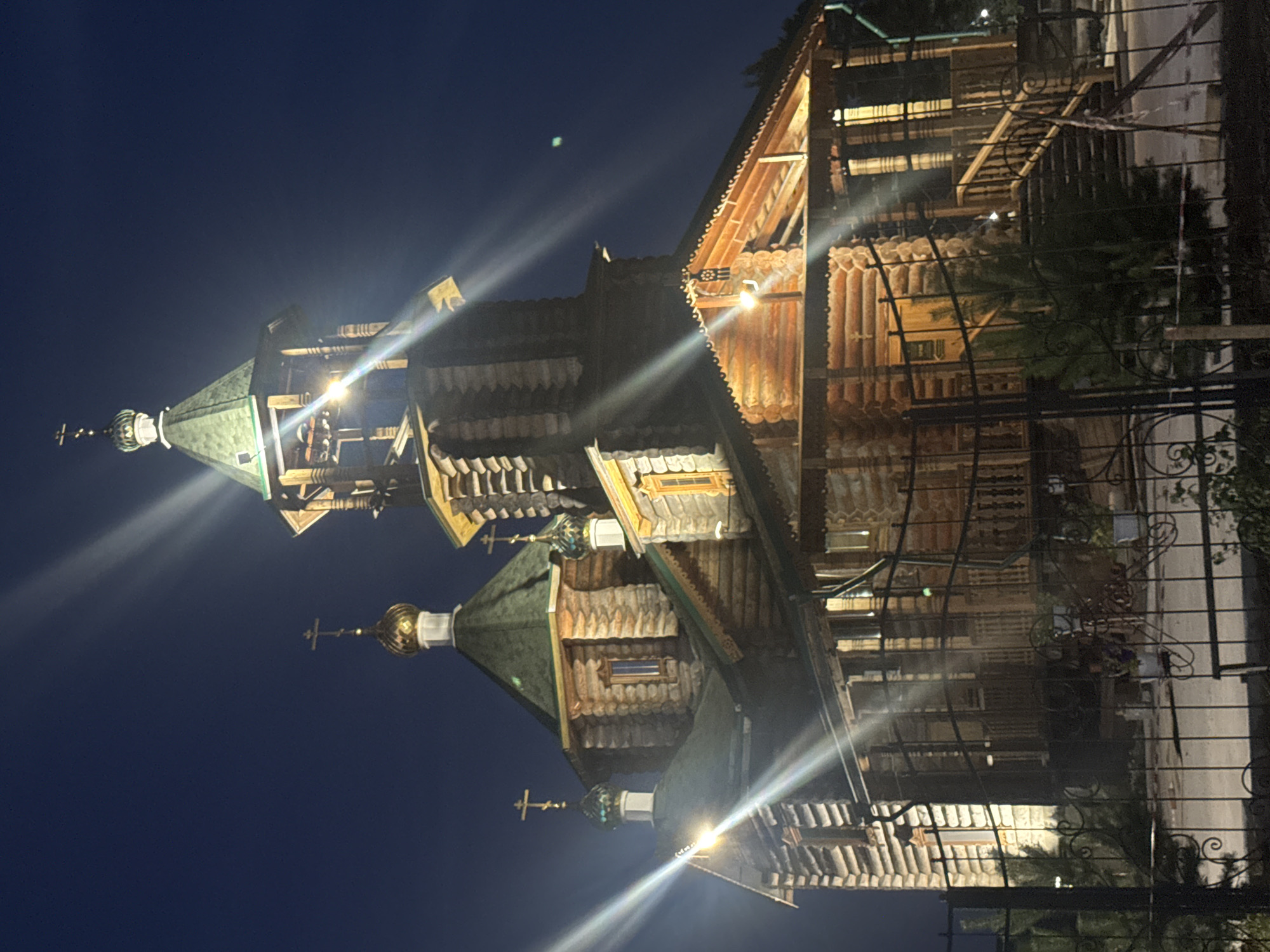 The image shows a wooden church at night, illuminated by artificial lights. The church has several domed roofs topped with crosses, and a bell tower. The architecture appears to be traditional Russian style. The church is surrounded by a fence, and some trees and plants are visible in the foreground. The sky is dark blue, indicating nighttime. Based on the architectural style, this is likely the Church of the Intercession of the Holy Virgin in Komsomolsk-on-Amur, Russia.