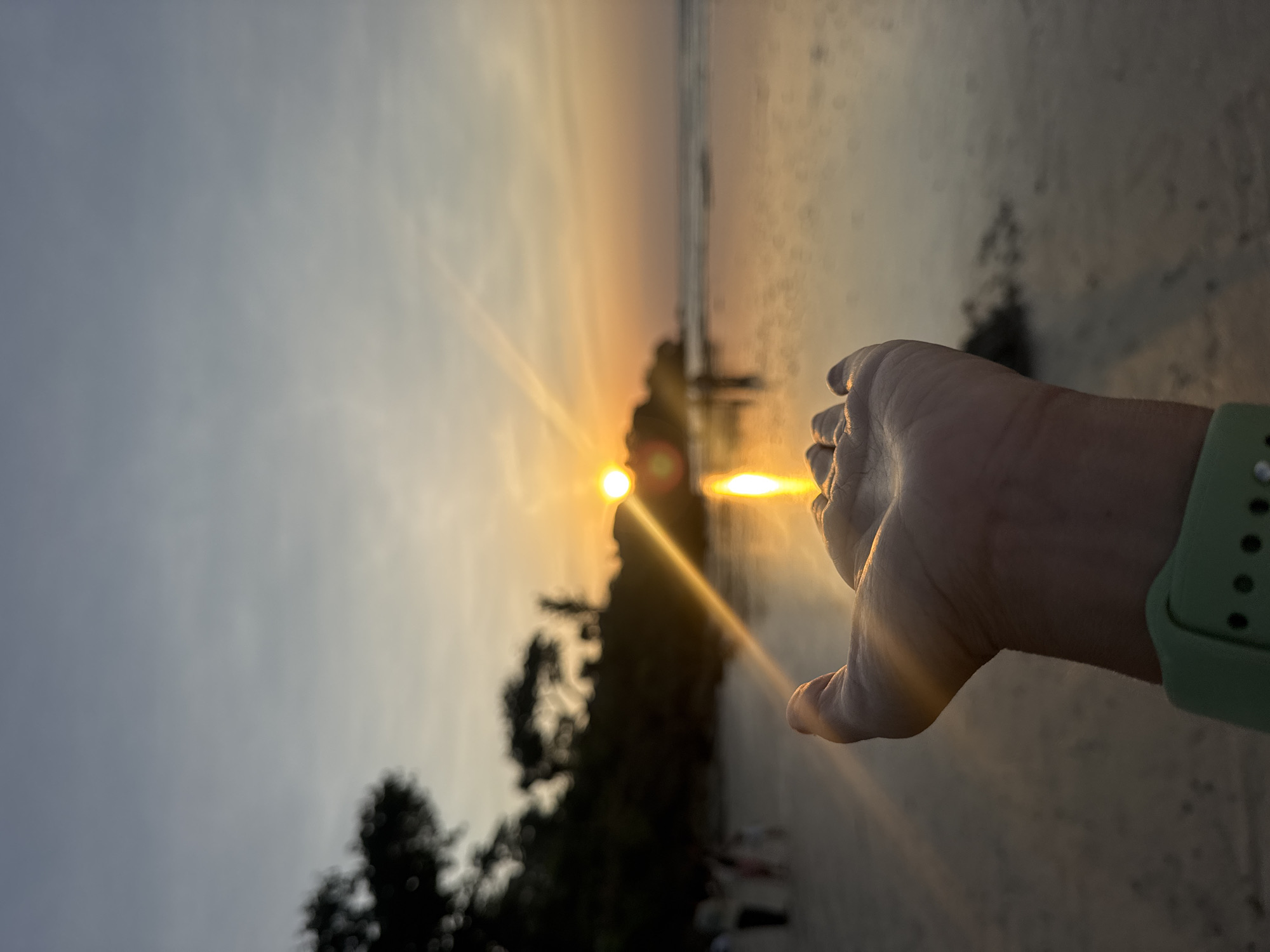 A close-up shot focuses on an open hand with its palm facing upward, positioned in the foreground. The hand appears to be cupping the setting sun, which is visible as a bright orb low on the horizon. Lens flares and light rays emanate from the sun, creating streaks of light across the image, particularly across the hand and reflecting on the water.
The background depicts a beach scene during sunset. The sun's warm golden and orange hues are reflected on the wet sand or shallow water that covers a large portion of the beach. Silhouetted trees and foliage are visible on a slightly elevated landmass to the left of the frame. Several out-of-focus figures, appearing to be people, are scattered along the beach in the mid-ground, suggesting a public space. The sky transitions from a pale blue at the top to warmer orange and pink tones closer to the horizon. The overall impression is a serene beach environment bathed in the warm light of a sunset. The watch band on the wrist of the person holding out their hand is a light green color. The location is Kuta Selatan, Indonesia.
