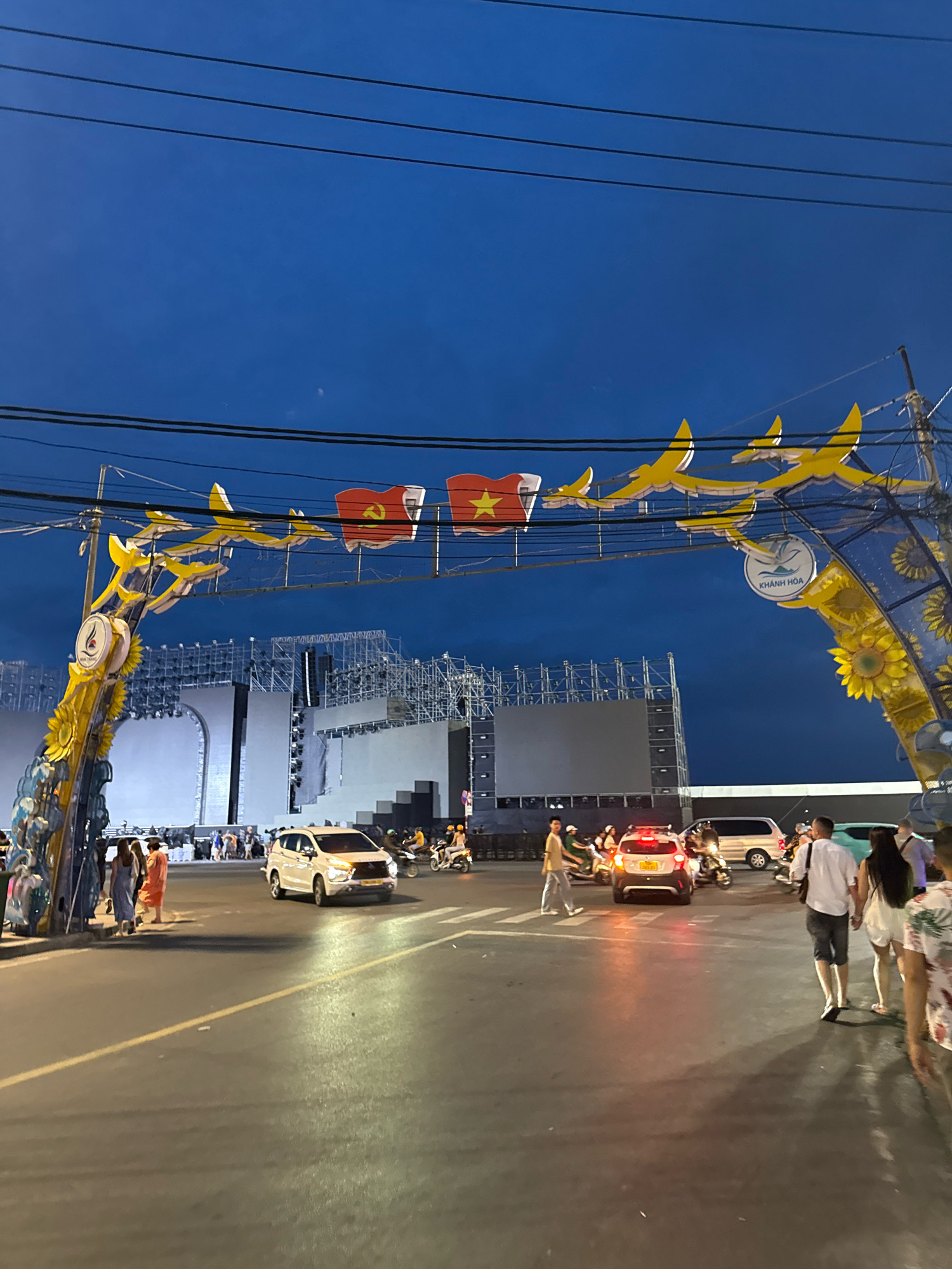 An outdoor archway decorated with yellow bird-like figures and sunflowers spans a street in Nha Trang, Vietnam, at dusk. Vietnamese flags are visible on the archway. Beyond the archway, a large stage setup with scaffolding and large screens suggests an event is taking place. Several vehicles, including cars and motorcycles, are on the street, and pedestrians are walking. The sky is a deep blue. The archway has a circular emblem with text that reads "KHÁNH HÒA".