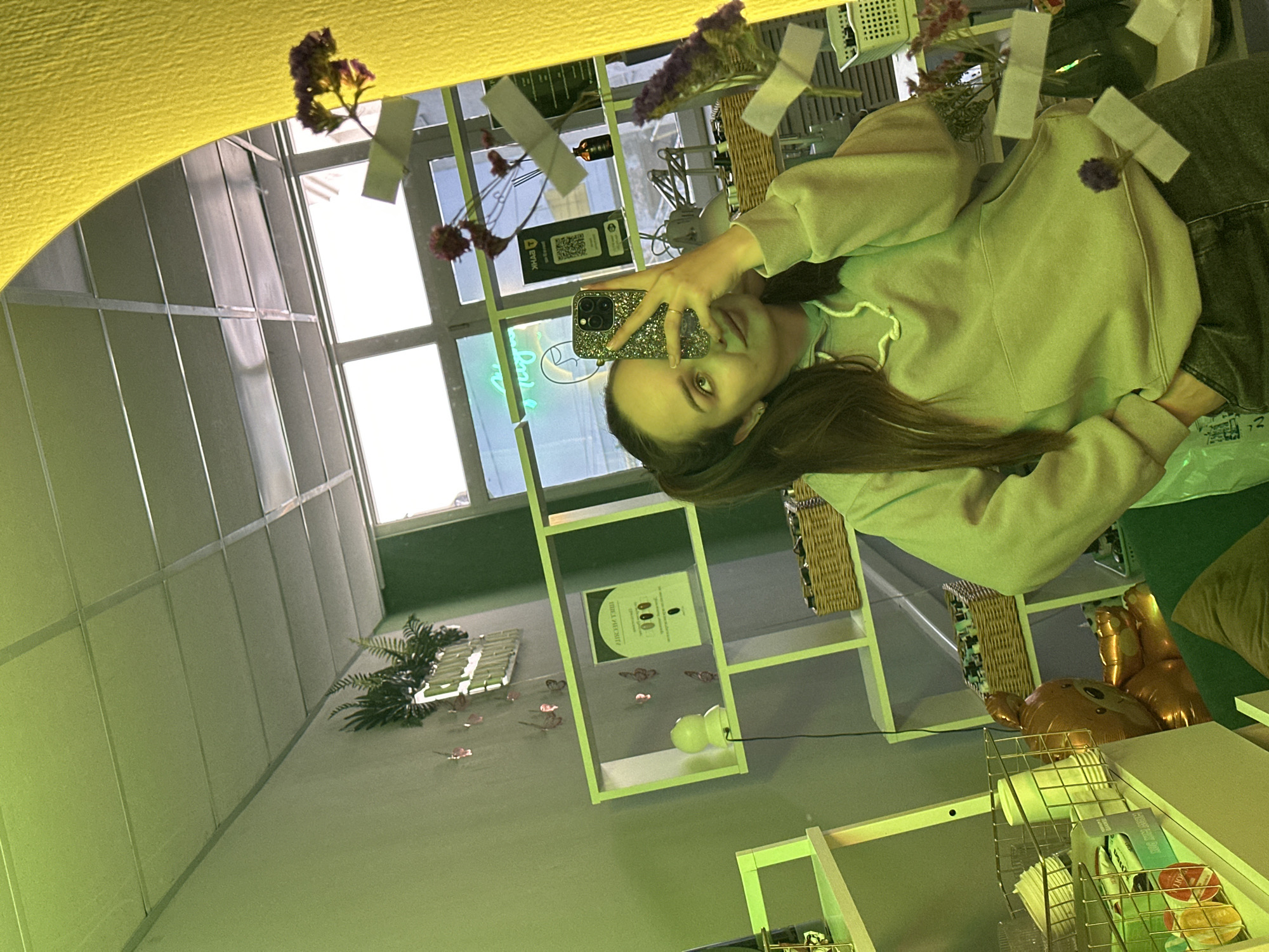 A woman is taking a selfie in a mirror. The reflection shows a room with shelving, plants, and decorations. Outside the window, there is a building. The selfie suggests the location is in a place with modern interior design, possibly a shop or a studio. There is no information to suggest it is specifically located in Chaikovsky, Russia.