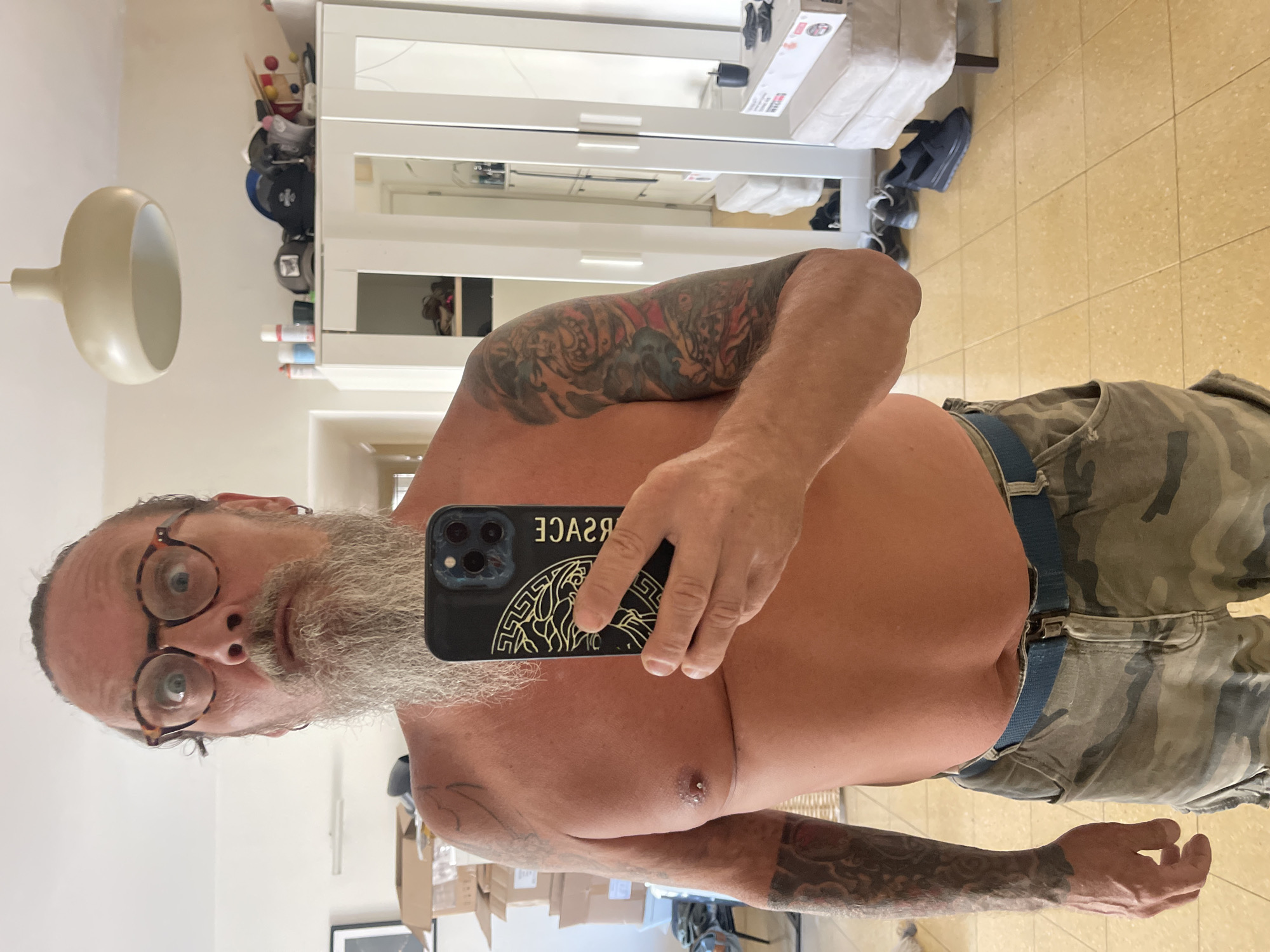 This media file is a horizontal, eye-level mirror selfie taken in an indoor setting, possibly a bedroom or dressing area, in Netanya, Israel. The primary subject is a man with a beard and glasses, shirtless and wearing camouflage pants with a blue belt. He has tattoos on his arms and chest, and a piercing on his chest. He is holding a smartphone in his right hand, which is angled to capture the reflection. The background features a white wardrobe with doors, a lamp on the left, and a tiled floor. Several objects are visible on the shelves of the wardrobe, including shoes and bottles. There is also a reflection of more of the room in the wardrobe doors, showing boxes stacked on a shelf. The lighting appears to be artificial, suggesting the photo was taken indoors, potentially during the day or evening. The man's expression is neutral. The text "VERSACE" is visible on the back of the smartphone case.
