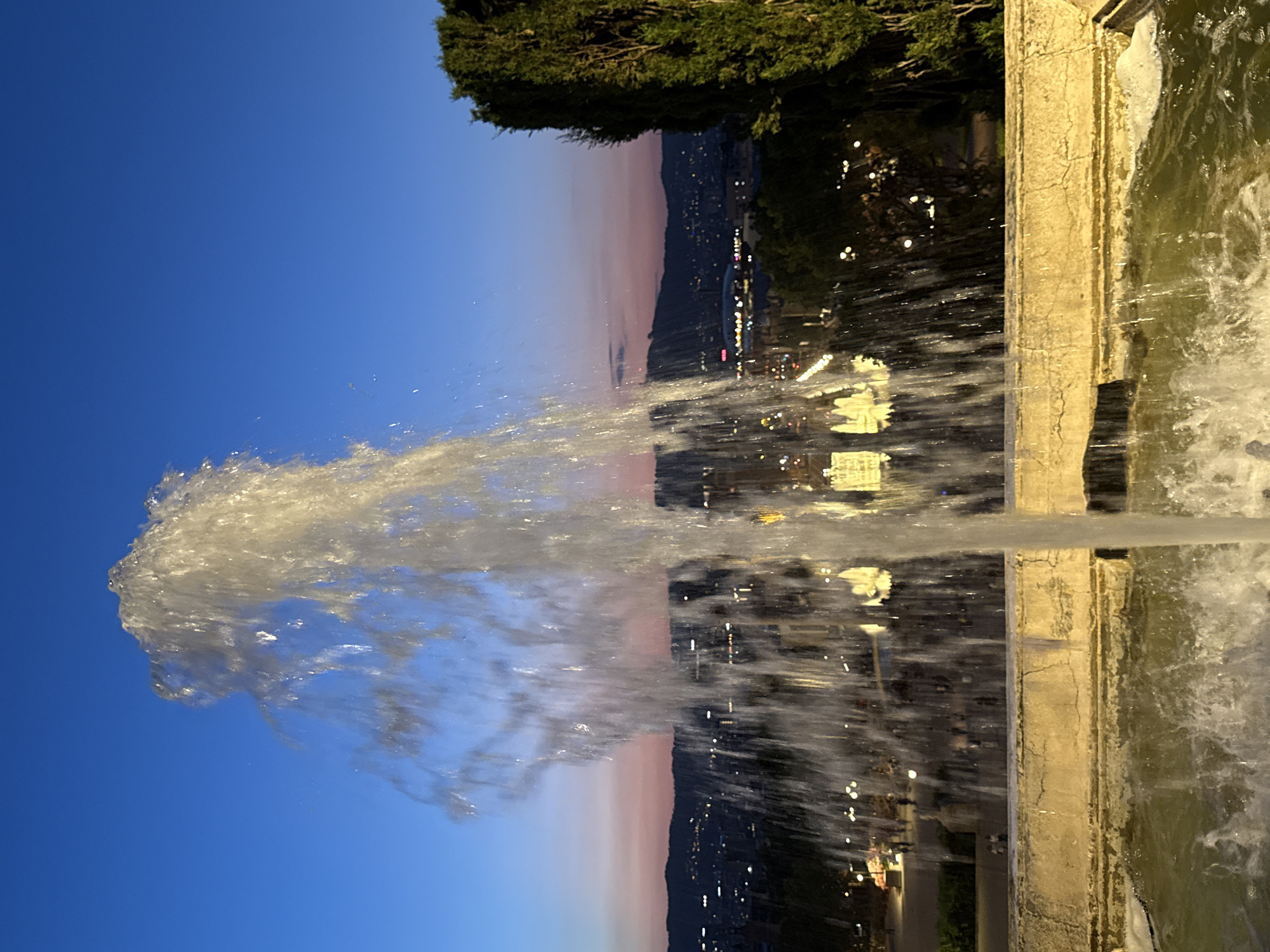 A water fountain is in the foreground, with water spraying upwards against a twilight sky. The fountain is made of stone and is filled with green water. In the background, the city of Barcelona, Spain, is visible with lights twinkling. The sky is a gradient of blue and pink, indicating it is dusk. There are trees on the right side of the image.
