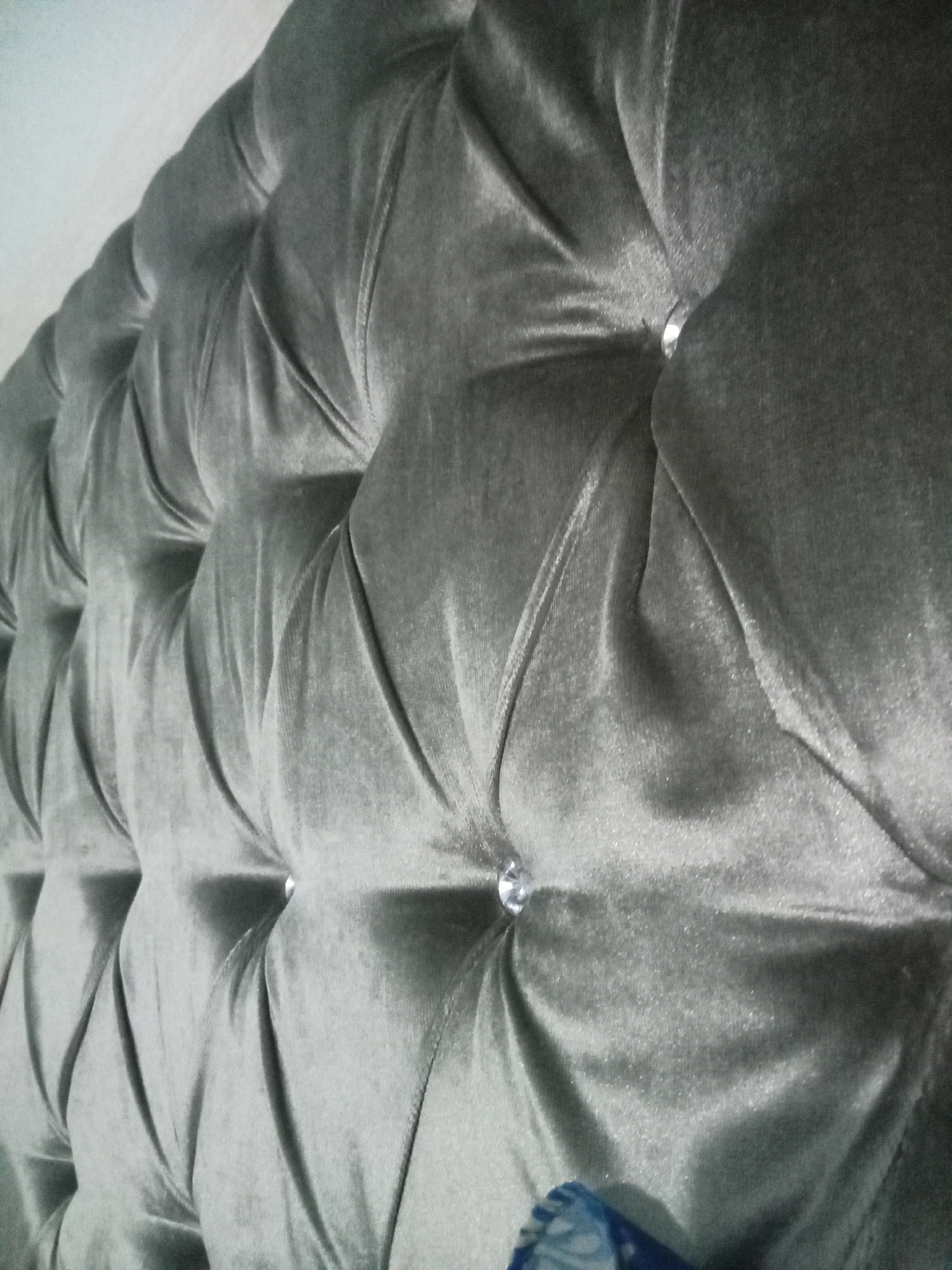 This image, captured in Baro, Indonesia, displays a close-up, angled view of a grey tufted upholstered surface, likely a headboard or sofa back. The material is a plush, velvety fabric, exhibiting variations in shade from light to dark grey due to light reflection and deep folds. The surface features a pattern of deep tufting, with multiple clear, possibly crystal or rhinestone, buttons embedded in the indentations, catching and reflecting light. The fabric gathers around these buttons, creating pronounced shadows and highlights that emphasize the textured surface. A plain, light-colored surface is visible in the upper left portion of the frame, serving as a background element. A small, obscured fragment of a blue and white patterned object is discernible at the bottom right corner. No individuals or specific actions are depicted.