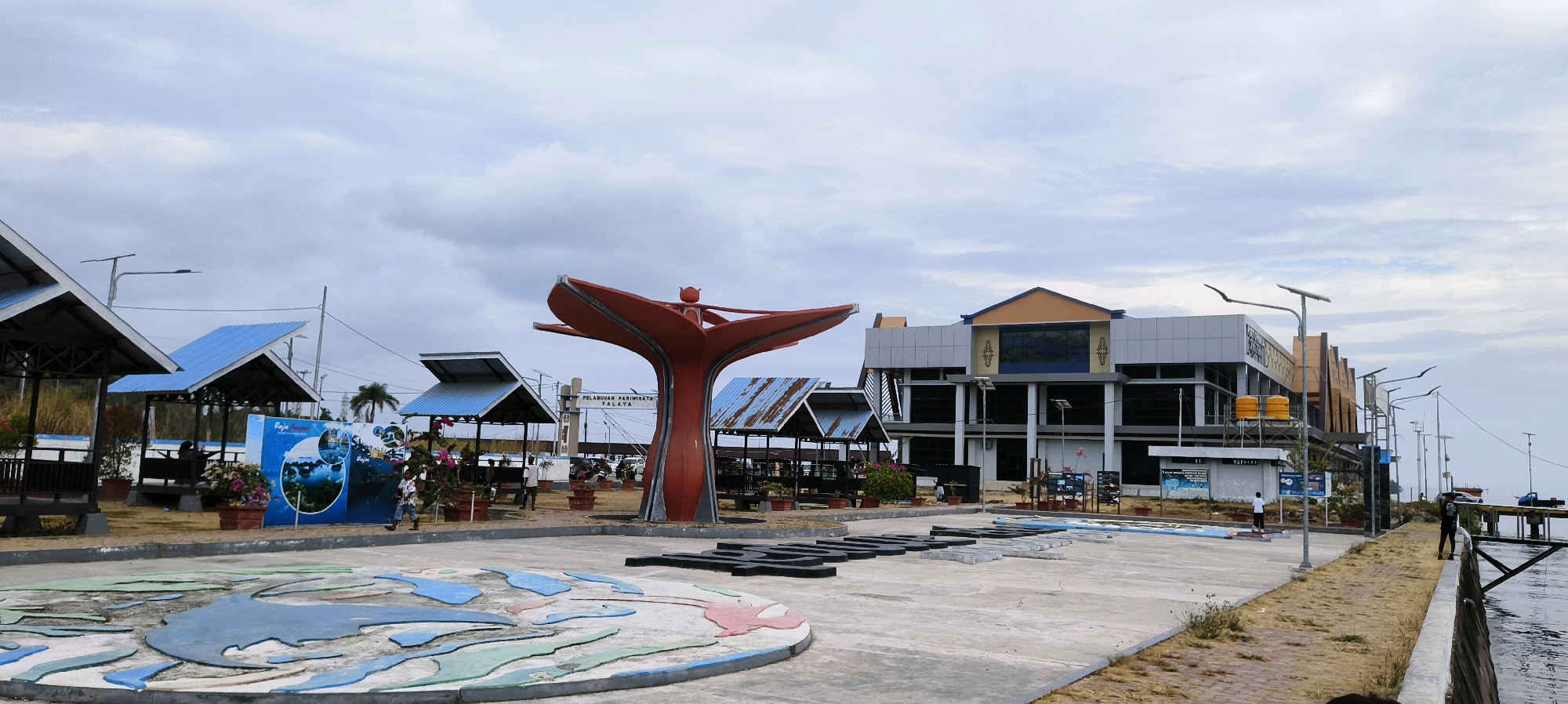 The image depicts a waterfront scene in the city of Waisai, Indonesia, likely a public recreational area or a port. In the foreground, a circular decorative element resembling a stylized map with blue and green landmasses is visible on a paved surface. Beyond this, a large, abstract orange sculpture that resembles a stylized flower or tree dominates the midground. To the left of the sculpture are several open-air shelters with blue roofs, and further back, a modern building with a gray facade and orange trim stands. Several people are present in the scene: two individuals are walking near the shelters, and one person stands near the waterfront on the right. The environment is characterized by a paved walkway, grassy areas, potted plants, streetlights, and water extending to the right of the frame, with a wooden pier visible. The sky is overcast with clouds.