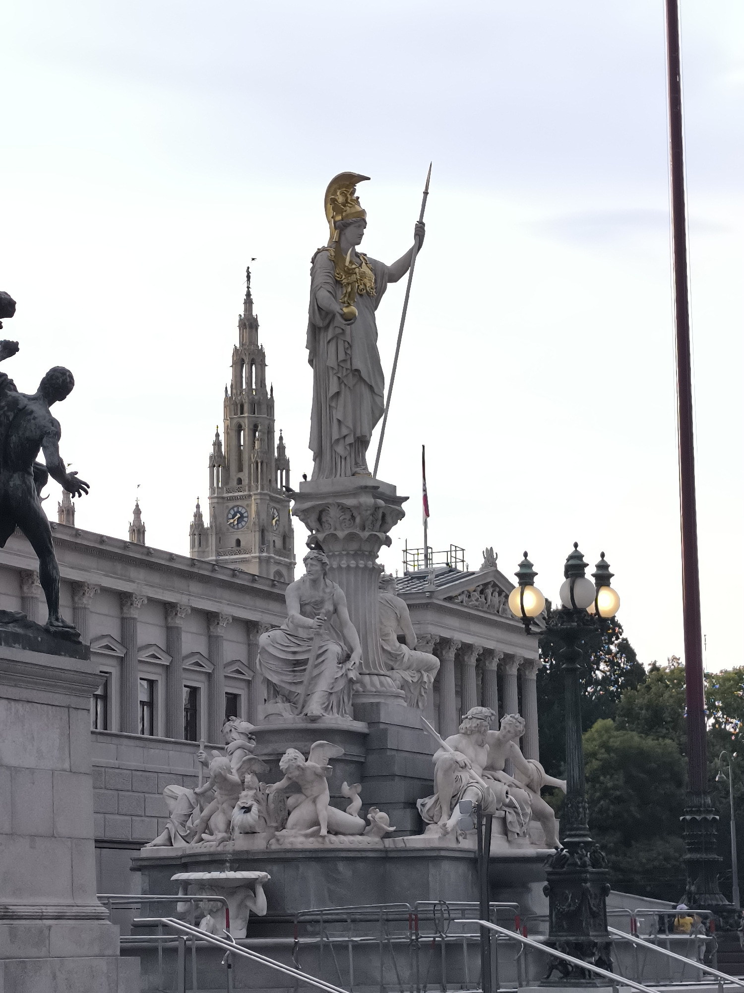 The image captures a view of the Pallas Athene statue in front of the Austrian Parliament Building in Vienna, Austria. The statue of Pallas Athene, adorned with a golden helmet and chest piece, stands prominently on a pedestal holding a spear. Around the base of the statue is a fountain with numerous smaller sculptures of figures and cherubs. The Austrian Parliament Building, with its classical architecture and columns, is visible behind the statue. Further in the background, a tall clock tower, part of the Vienna City Hall (Rathaus), stands against a pale sky. Several lampposts with illuminated globes are positioned to the right of the main statue. The scene appears to be set outdoors, with some trees visible behind the buildings. There are no visible people directly interacting with the statues or buildings, though some individuals may be present in the distance. The overall impression is one of a grand public space and a significant historical landmark.
