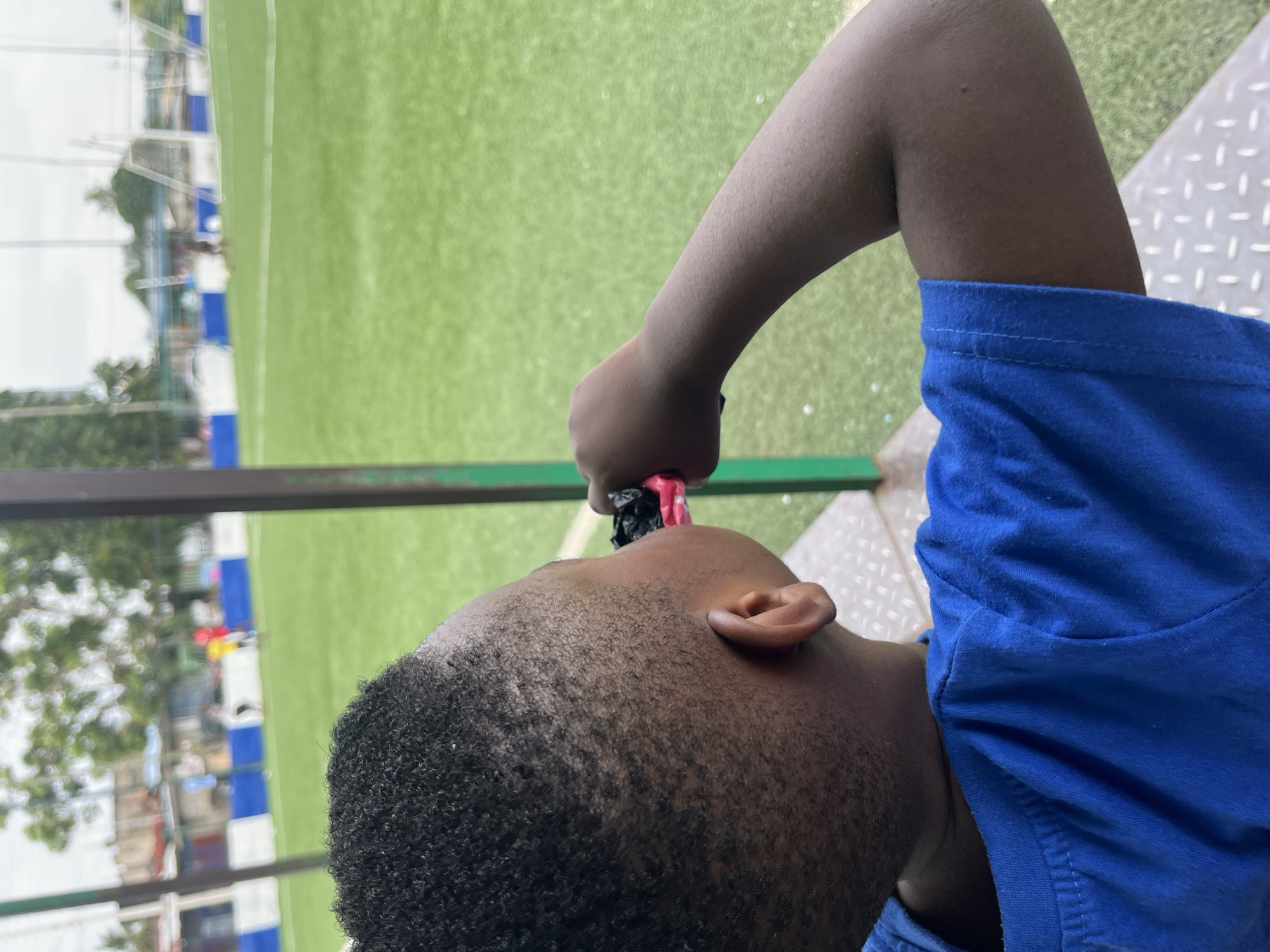 In Accra, Ghana, a young boy wearing a blue t-shirt is seen from behind, looking towards a bright green artificial turf soccer field. He has a short haircut and his right arm is raised, bringing a pink and black object to his mouth. The field has white markings and goalposts are visible in the distance. A blue and white fence borders the field, and behind it, buildings and trees can be seen. The sky is overcast. The boy is sitting on a textured surface, possibly a bench or bleacher.