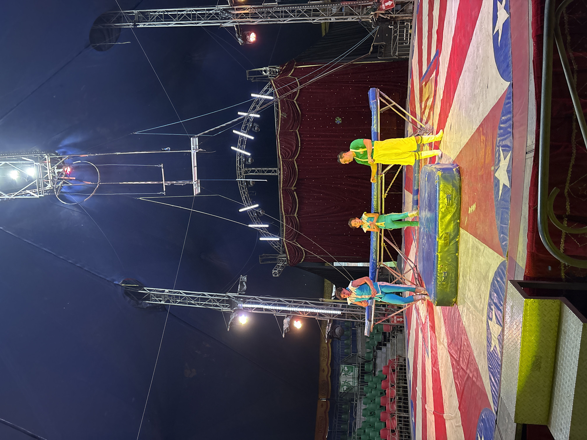 The image depicts a circus performance in what appears to be a big top tent. Three performers are visible on a raised platform with an American flag-themed backdrop. One performer is dressed in yellow and green, another in green and teal, and the third in blue and teal. The tent has a blue ceiling with rigging and lighting equipment suspended from it. There are empty seats visible in the background. It is in Tejalpa, Mexico.