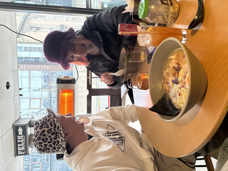 Two individuals are seated at a light brown, round wooden table inside Felix Bar & Kitchen, located in Belgrade, Serbia, visible through a large window overlooking an urban street. On the left, a person with dark hair, wearing a white leopard-print bucket hat and a white hoodie with a black geometric logo, smiles while looking towards the second individual. On the right, a person with pink-tinted hair, wearing a dark purple fuzzy bucket hat and a black puffer jacket, is actively eating ramen from a light-colored bowl using a white ceramic spoon. A second bowl of ramen is in the foreground, containing noodles, fried protein, lime slices, red spice flakes, purple edible flowers, and a piece of nori seaweed. Drinks on the table include a clear stemmed glass with an amber liquid, a tall clear glass with an orange beverage and ice containing a red straw, a red Coca-Cola can, and another clear glass with a lime wedge. A glass jar with a green lid, filled with spices, and a black ceramic spoon are also on the table. An operational infrared heating lamp, emitting an orange glow, is visible mounted on the ceiling outside the window.