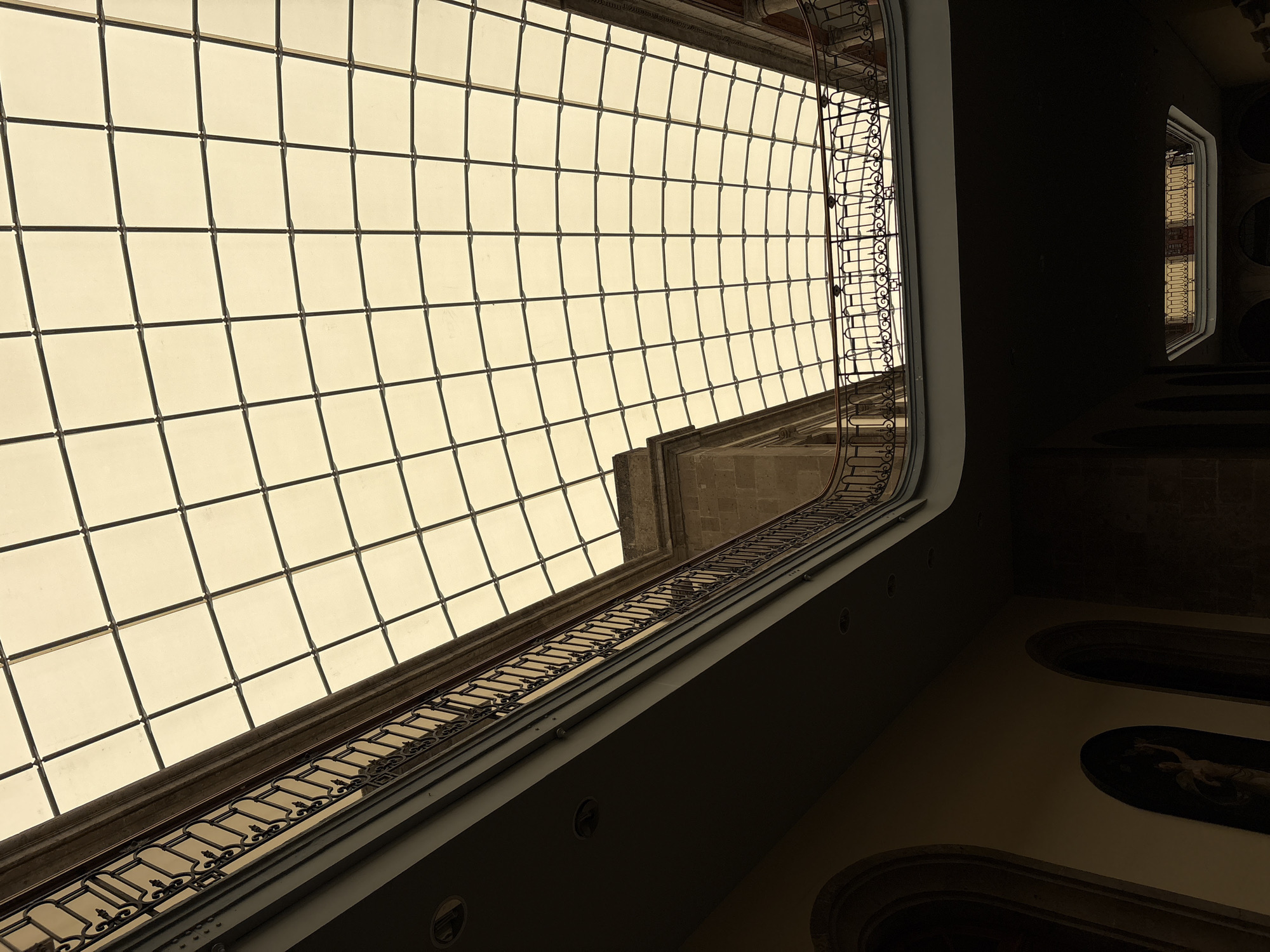 View of the interior of a building in Mexico City, Mexico. The most prominent feature is a large, rectangular skylight composed of numerous small square panels. Dark metal supports form a grid pattern across the skylight.
Beneath the skylight, ornate metal railings are visible along what appears to be upper floors or walkways within the building. The walls are painted a dark color. Two paintings in rounded frames are visible.