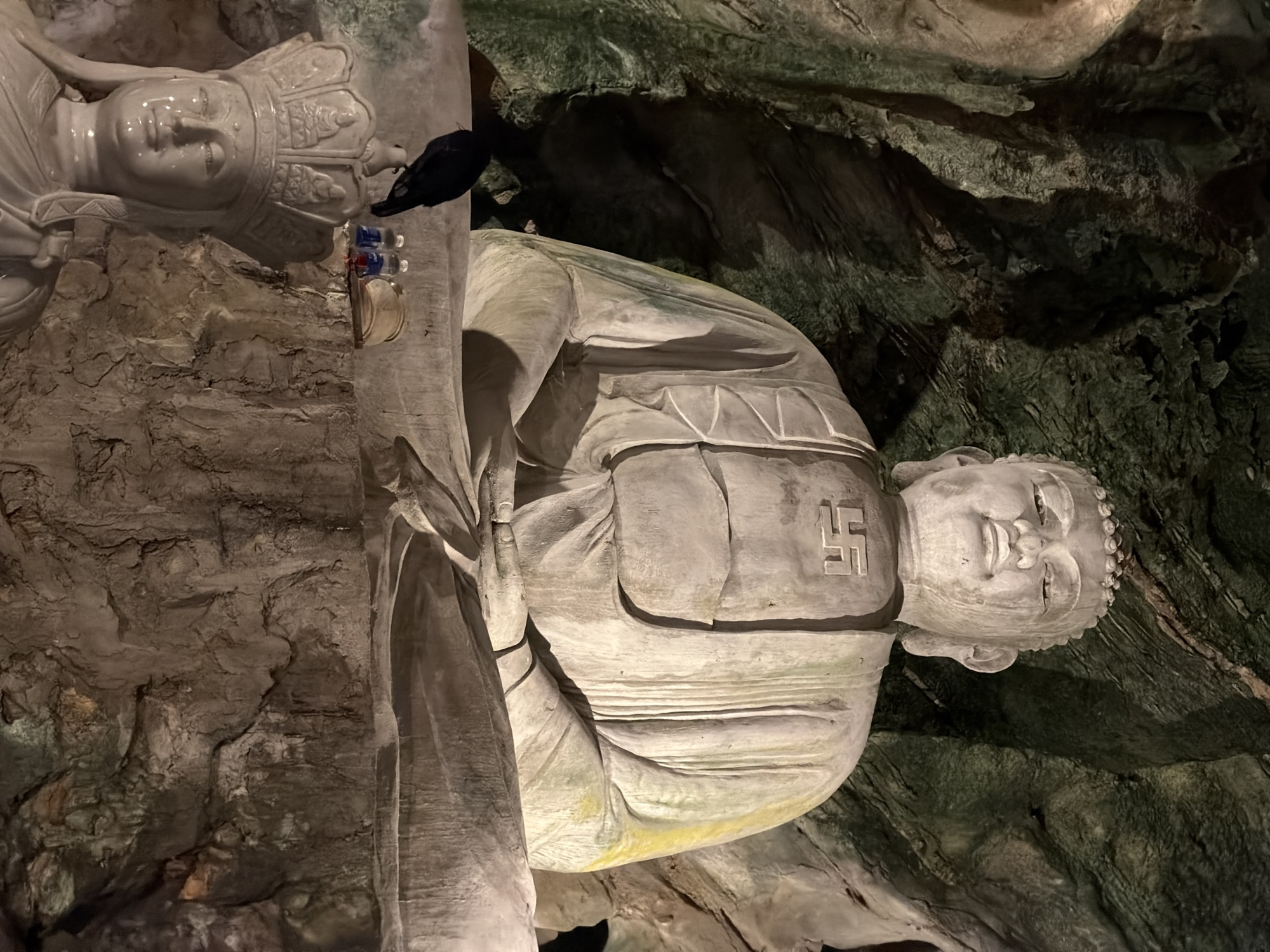 The image shows a stone statue of Buddha inside a cave, possibly at the Marble Mountains in Đà Nẵng, Vietnam. The Buddha is adorned with a swastika symbol on its chest. A smaller statue with a crown is situated above it, along with some small colorful bottles. The background consists of rough cave walls.