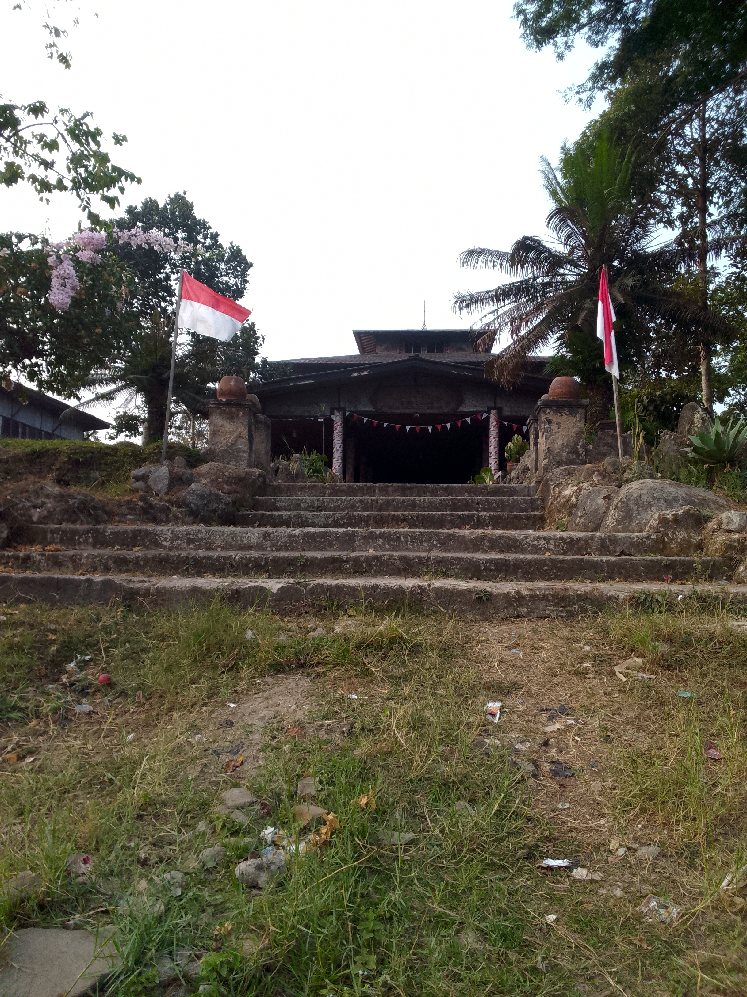 The image captures an outdoor scene in Padalarang, Indonesia, featuring a weathered stone staircase leading up to an old, traditional building. The building has a dark, sloping roof and is adorned with a string of small red and white triangular flags, suggesting a festive or commemorative occasion. Two Indonesian flags, red and white, are prominently displayed on flagpoles flanking the entrance to the building. The building appears to be constructed with natural materials, possibly stone and wood, and has a somewhat dilapidated, aged appearance. Surrounding the structure are lush green trees, including a palm tree on the right, and a tree with pale pink flowers on the left. The ground in the foreground is covered with overgrown grass and scattered debris, indicating a lack of recent maintenance in that area. The sky is overcast, casting a soft, diffused light over the scene. The overall impression is one of historical significance and a sense of local culture, possibly a place of worship or a community gathering spot.