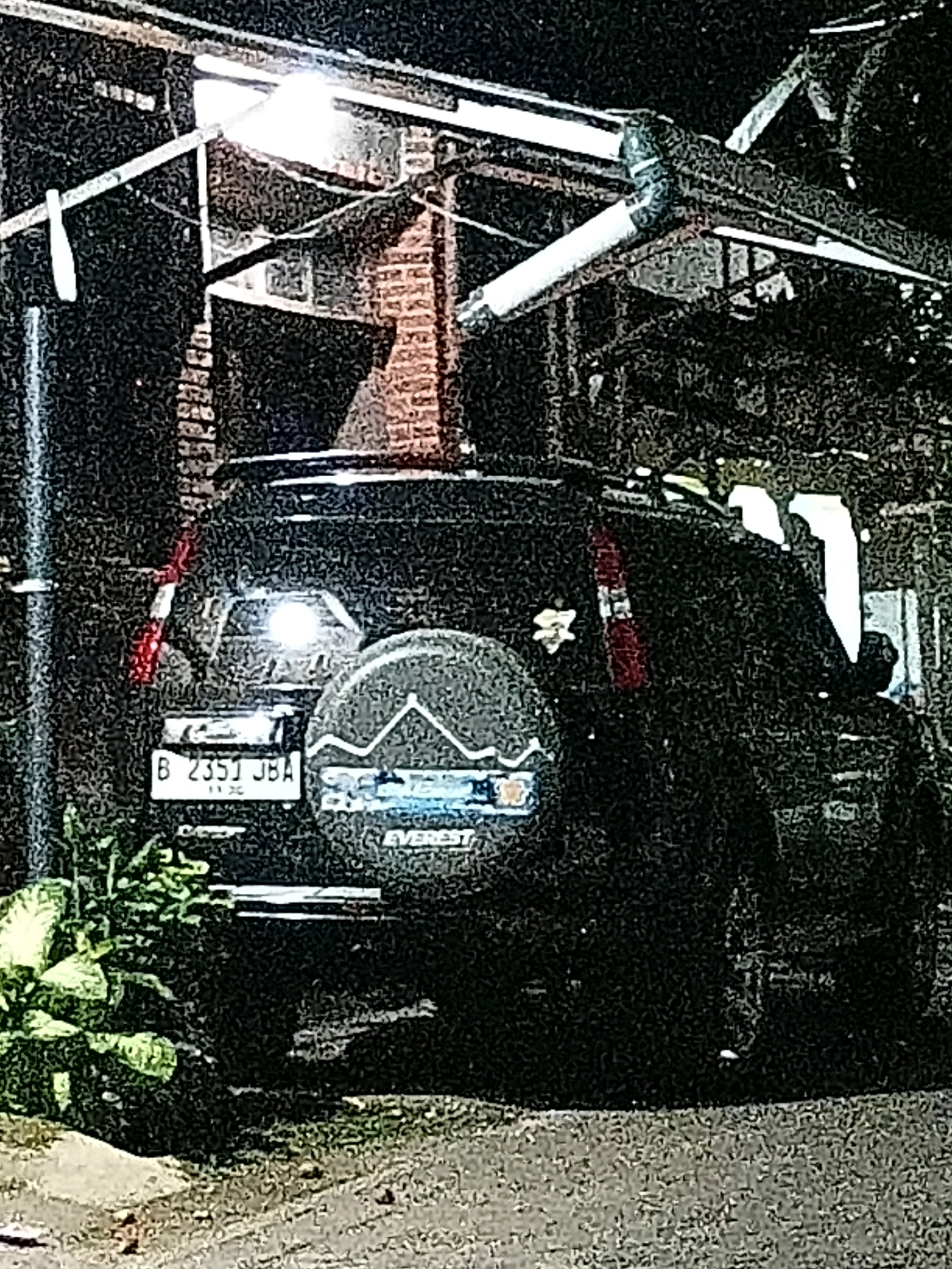 A black SUV with a spare tire mounted on the back is parked on a paved surface near some vegetation. The SUV has a license plate that reads "B 2351 JBA". Behind the SUV, there is a brick structure with a metal overhang and some lighting. The scene appears to be outdoors at night, possibly in an urban or semi-urban area. There are no people visible in the image.