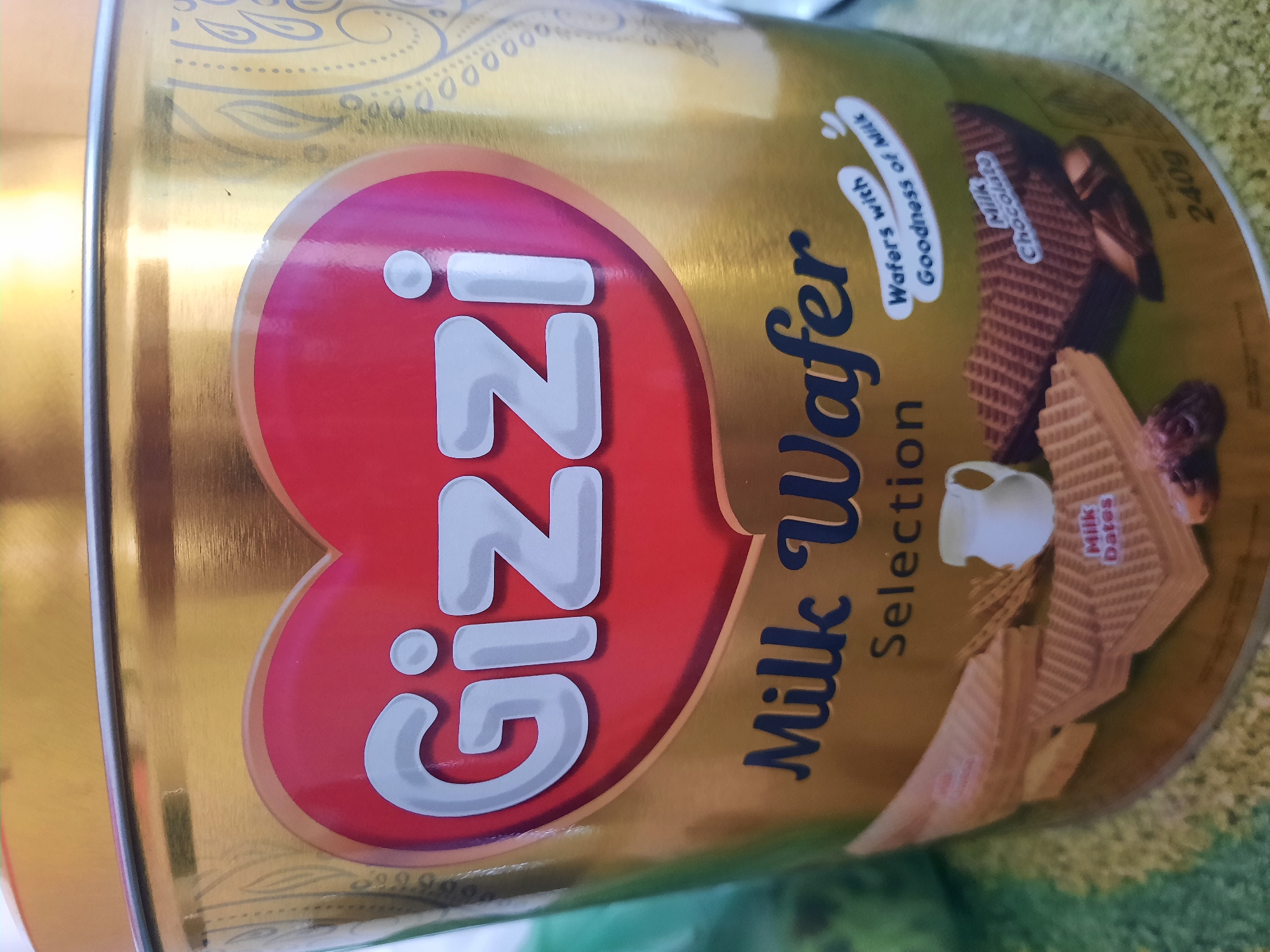 A close-up, eye-level shot shows a gold-colored tin can for "Gizzi Milk Wafer Selection." The can features a large red heart with the "Gizzi" logo in white, bubbly letters. Below the logo, "Milk Wafer Selection" is written in a dark blue, cursive font. Various wafers are depicted at the bottom, including one labeled "Milk Dates" and another labeled "Milk Chocolate." A small white jug is also visible. The background is blurred and appears to be an indoor setting. The can is likely displayed in a shop or home environment within Lhokseumawe, Indonesia.