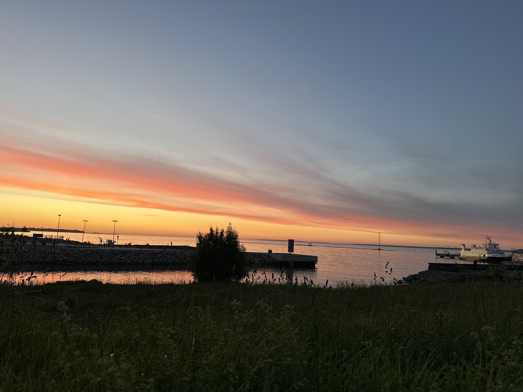 A wide, low-angle shot shows a tranquil coastal scene in Tallinn, Estonia during a sunset. The sky is a gradient of colors, transitioning from a soft orange and pink near the horizon to a pale blue higher up, with thin clouds stretching across the sky. The calm sea reflects the vibrant sky colors. In the foreground, tall green grass and reeds are silhouetted against the brighter background. Along the waterfront, a breakwater made of large rocks separates the sea from a paved area. Several lamp posts with lights on are visible on the paved area, along with benches and playground equipment. In the distance, a white ferry is docked at a pier. A few figures can be seen on the waterfront and on the ferry. The overall impression is one of peaceful evening serenity.