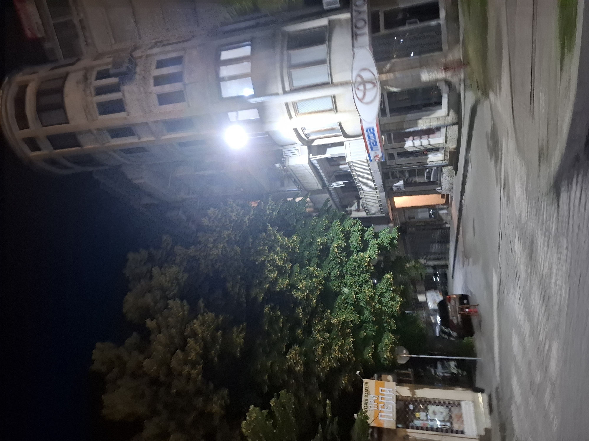 The image depicts a street scene at night. On the right side of the frame, a multi-story building with a curved facade and several windows is visible. A sign with the "Toyota" logo and another sign that reads "Mazda" are prominently displayed on the ground floor of this building. To the left of this building, a large tree with dark green foliage is present, obscuring part of the building and the street. In front of the tree, a storefront with a sign reading "ПОДАРЪЧЕН МАГАЗИН ПРИ ПЕПА" (Gift Shop At Pepa) is visible. The street is paved with cobblestones, and a car is parked on the side of the road. The lighting suggests it is nighttime, with streetlights illuminating the scene.