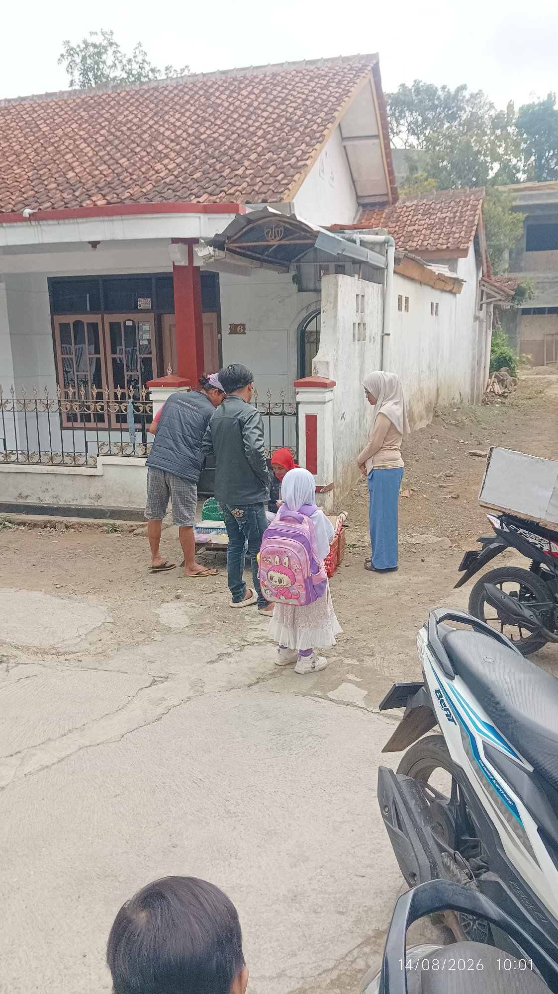 The image shows a group of people standing outside a house with a tiled roof. The house appears to be in a residential area. In the foreground, a young girl wearing a white dress and a pink backpack with cartoon characters is standing. To her left, two men in casual clothing are talking. Another person in a red headscarf is partially visible. To the right, a woman in a hijab and blue pants is looking away. A motorcycle with a white and blue design is parked on the right side of the image. Another motorcycle is partially visible in the bottom right corner. The ground is paved with concrete. The timestamp "14/08/2026 10:01" is visible in the bottom right corner. The overall scene suggests a gathering or interaction in front of a home.