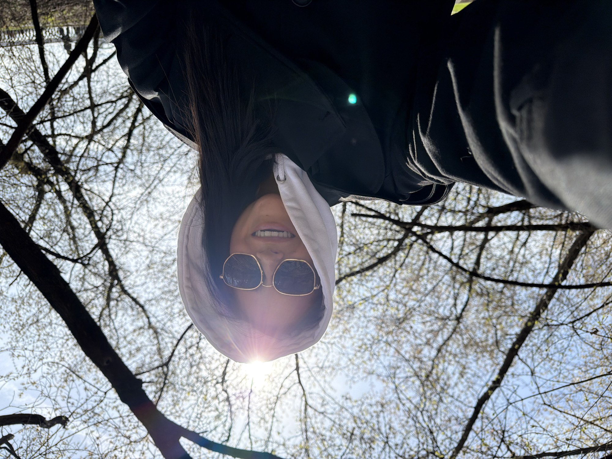 A woman wearing sunglasses and a white hooded sweatshirt is pictured from a low angle against a backdrop of bare tree branches and a bright sky. The sun is directly behind her hood, creating a lens flare. She is looking down at the camera and smiling, revealing her teeth. She is wearing a black jacket. The location is in a park in Khimki, Russia.