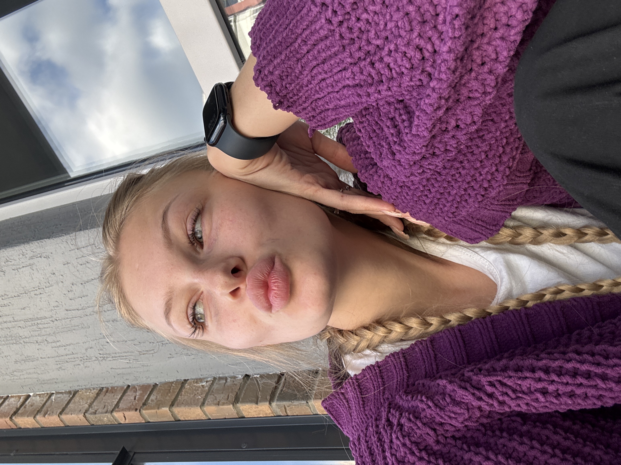 In this portrait, a young woman with blonde hair, styled in two braids, poses with her hand propping up her head. She is wearing a thick, knitted purple cardigan over a white t-shirt and dark pants. A black smartwatch is visible on her left wrist. The background suggests an urban setting, with a textured grey wall and a window reflecting a cloudy sky. A brick column is visible on the left side of the frame, hinting at an outdoor or semi-outdoor location, possibly a balcony or patio in the city of Perm, Russia. The woman's expression is playful, with her lips puckered as if blowing a kiss, and she is looking off to the side.