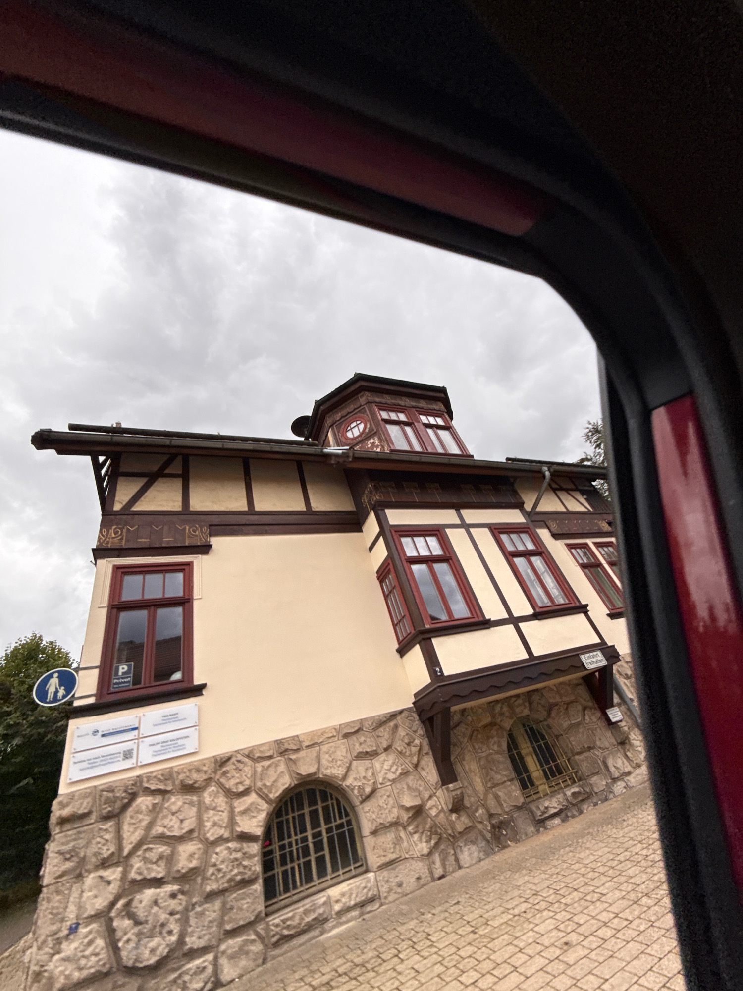 This image, taken from inside a vehicle in Bad Sachsa, Germany, captures a unique building with a cream-colored facade and dark brown timber framing. The building has multiple red-framed windows, some of which are bay windows extending from the upper floors. The ground floor is constructed with large, rough-hewn stones, and features an arched window with a metal grate. A paved walkway, made of light-colored rectangular stones, leads up to the building. The sky is overcast with gray clouds. Signs are visible on the building, including a blue circular sign with a white silhouette of two people, one adult and one child, and a parking sign with a "P" and the word "Privat". The framing of the image suggests it was taken through a car window, with the dark interior of the vehicle visible along the edges.