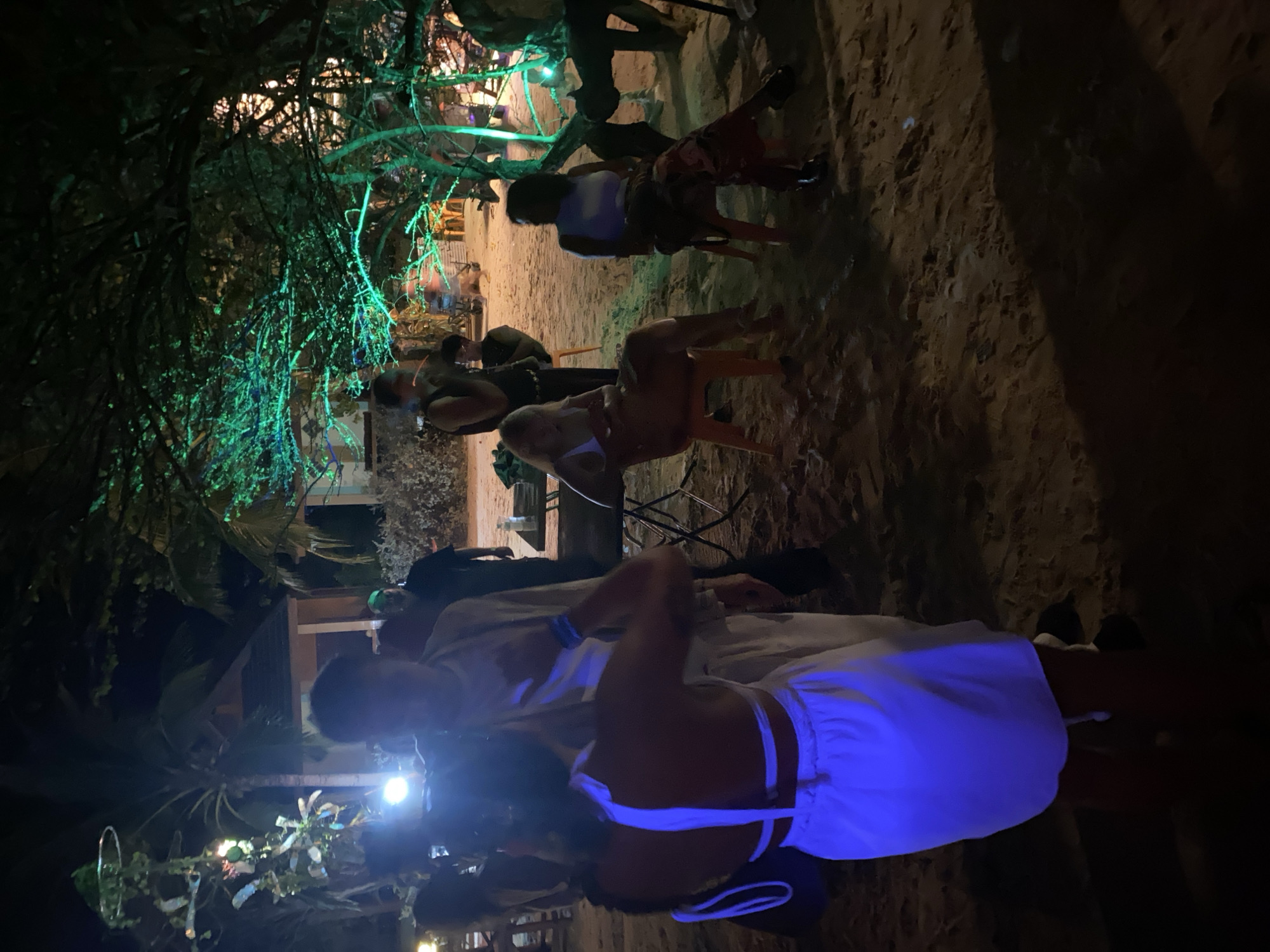 The scene depicts an outdoor gathering at night, likely on a beach. The location is in ตำบลเกาะพะงัน, Thailand. There are multiple people present, some sitting on chairs and others standing. They appear to be engaged in conversation and socializing. Artificial lighting is visible, including green lights on trees. The environment includes trees, sand, and what appears to be a building in the background.