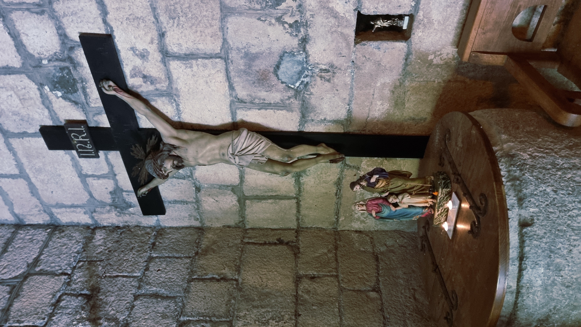 The image shows a religious altar within a stone-walled room. A large black crucifix with a statue of Jesus Christ hangs on the wall. Below the crucifix, on a circular wooden table, are two lit candles and a statue of the Holy Family, depicting Mary, Joseph, and the infant Jesus. To the right of the crucifix, a small niche in the wall contains a miniature statue of a saint. A wooden bench is visible to the right of the altar. This scene is located in the cloister of the Monastery of Sant Pere de Camprodon, in Camprodon, Spain.
