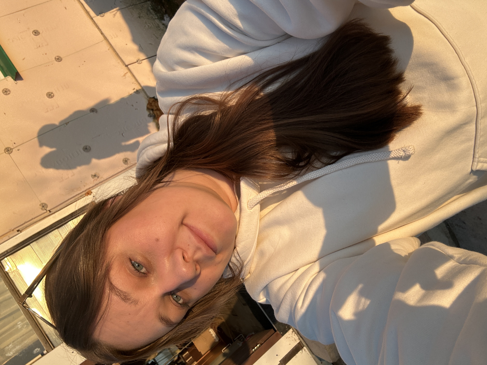 A woman with brown hair and blue eyes is wearing a white hoodie. The photo appears to be taken outside, possibly on a roof, with sunlight illuminating the scene. The location is not identifiable as being in the city of Ольховка, Russia, based on the visual information provided. The background shows a building with exposed insulation panels and a window reflecting sunlight.