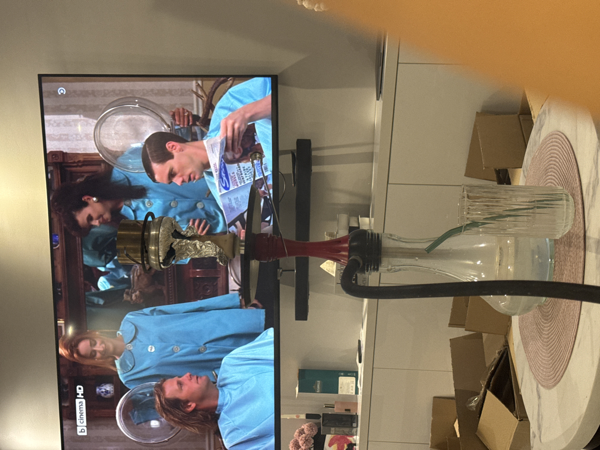 The image depicts an indoor scene with a television screen displaying a film, and in the foreground, a hookah setup. The television shows a scene from a movie or TV show featuring three individuals. Two women, dressed in teal garments, appear to be operating salon-like equipment, with one woman in the background and another in the mid-ground. A man is seated in the foreground of the TV screen, wearing a teal smock and holding a magazine.
In front of the television, a red and clear glass hookah with a black hose is positioned on a table. Beside the hookah is a clear glass with a green straw. The table itself is round with a marble-like surface and has a pink, circular placemat. In the background, on a shelf below the television, are various items including a green bottle with "Bushmills" branding, smaller bottles, and some cardboard boxes. The overall environment appears to be a domestic setting. The image does not contain any specific indicators to determine its location within Burgas, Bulgaria.